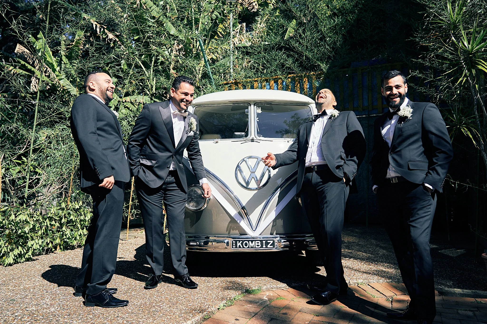 Four men in tuxedos standing in front of a vintage white Volkswagen van, enjoying a moment of laughter and camaraderie outdoors with green foliage background.