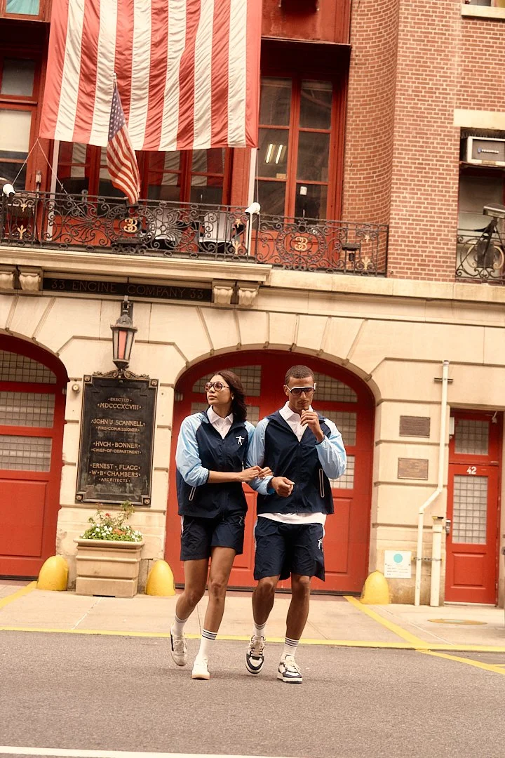 Two young people walking in front of a historic building with a large American flag hanging from the balcony. The woman has long dark hair, glasses, a navy vest, white shirt, navy shorts, and white sneakers. The man has short hair, sunglasses, a navy