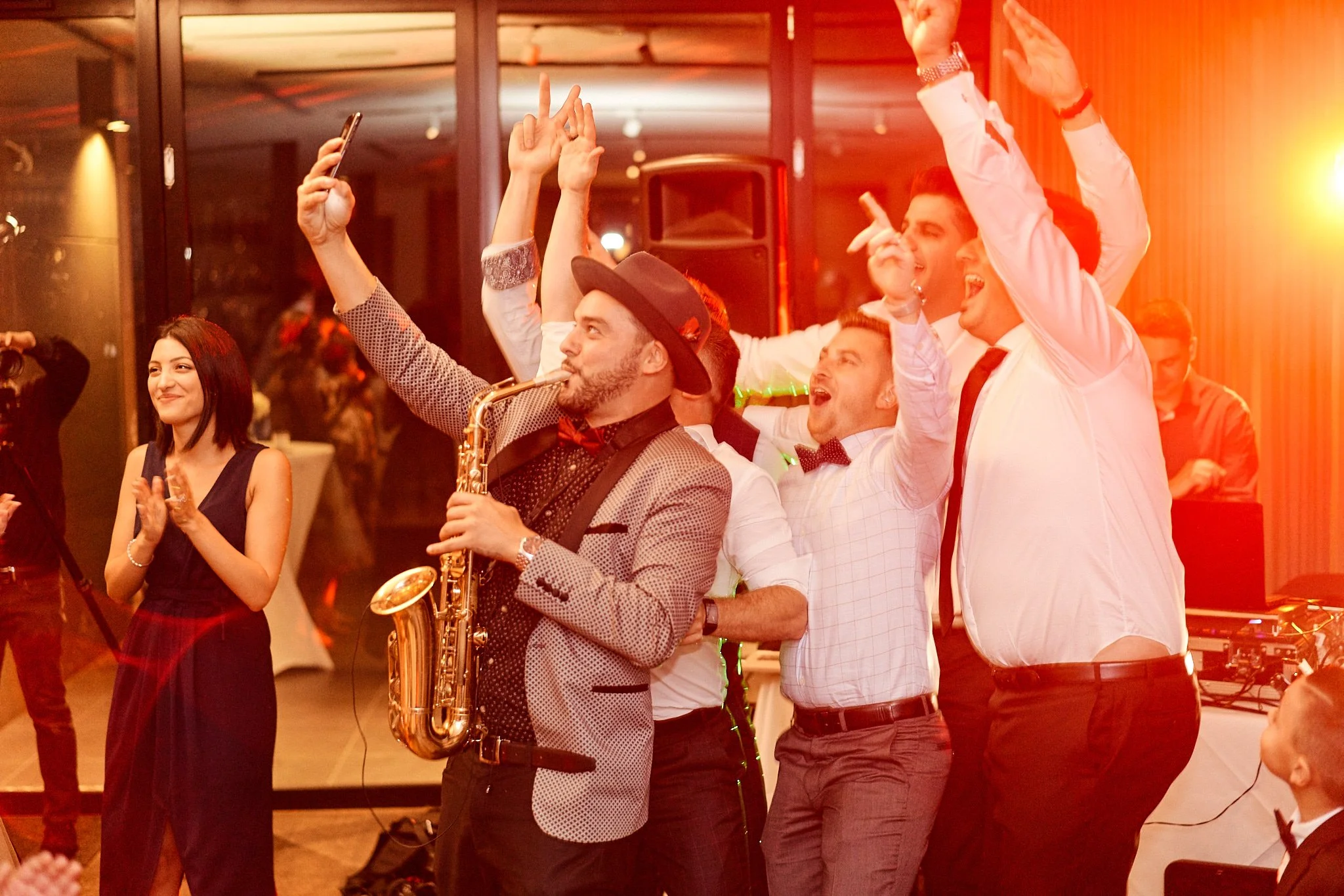 Group of people dancing and celebrating at a party, with a man playing the saxophone in the foreground.