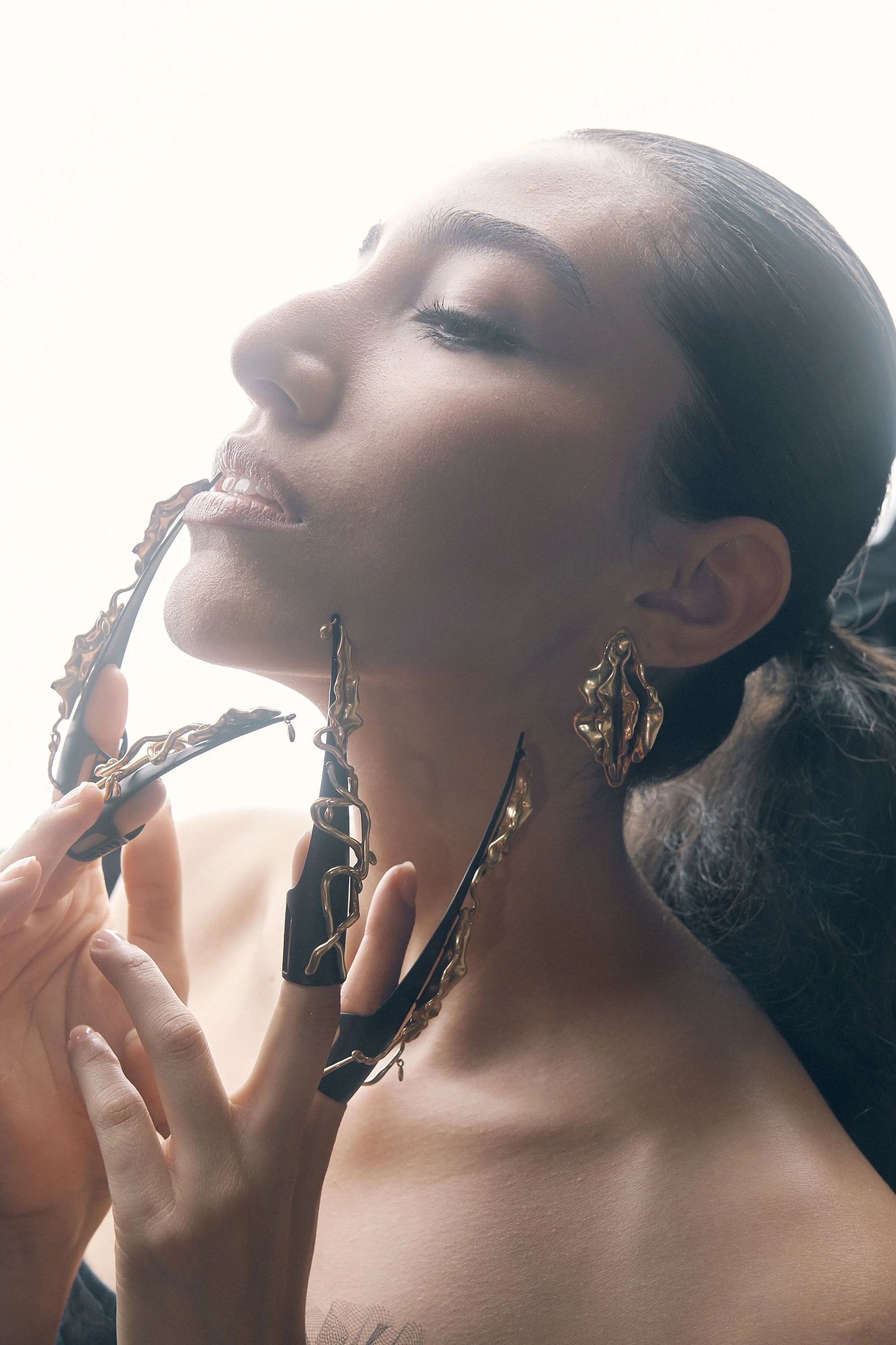 A woman with dark hair styled in a slicked-back manner, holding a pair of glasses decorated with gold and black accents near her face. She has a serene and confident expression, with makeup emphasizing her eyes, lips, and cheekbones, and wears large,