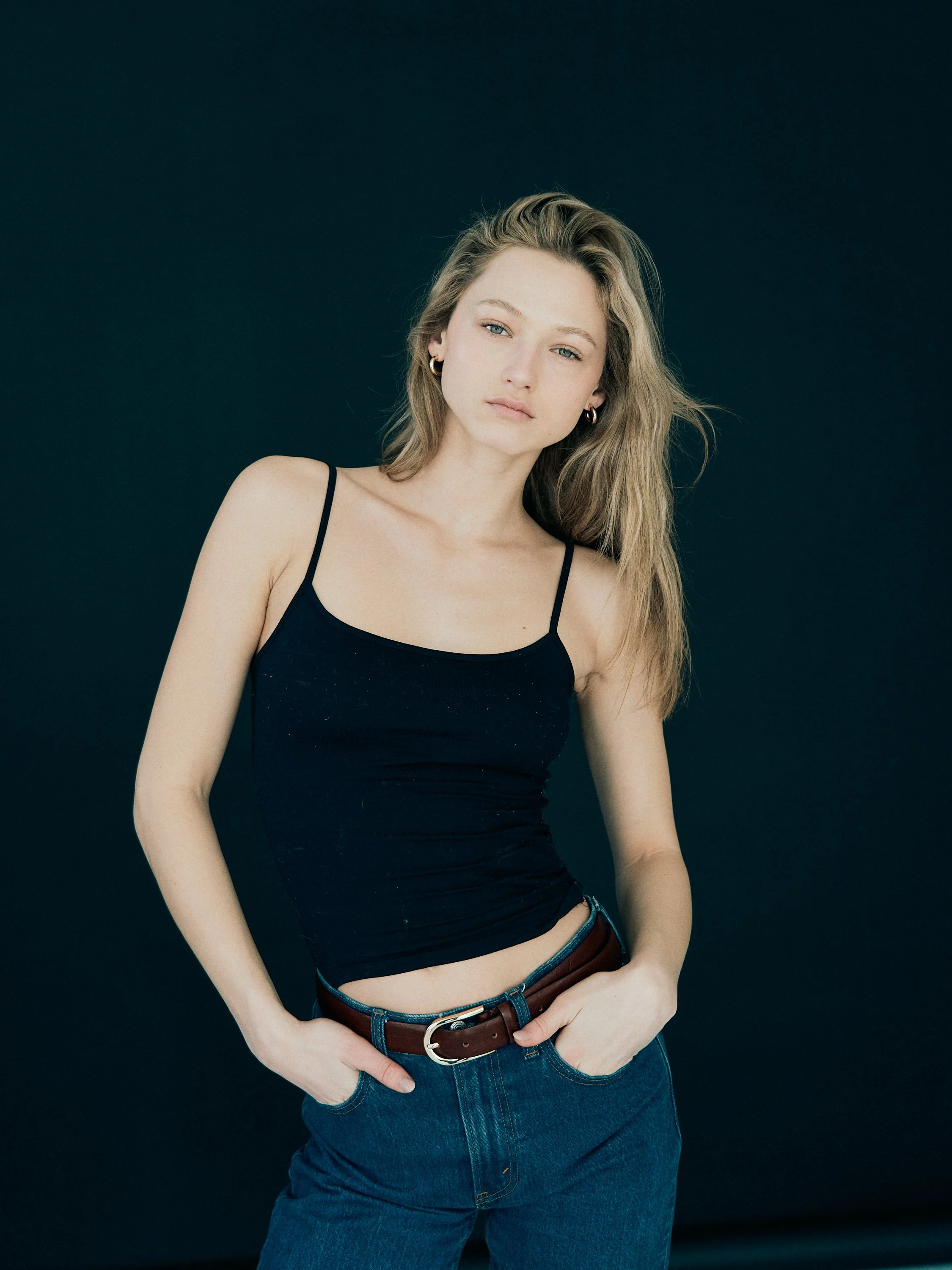 A young woman with long, wavy blonde hair and fair skin posing against a dark background. She is wearing a black spaghetti strap top and blue jeans with a brown belt, with her right hand in her pocket and looking toward the camera.