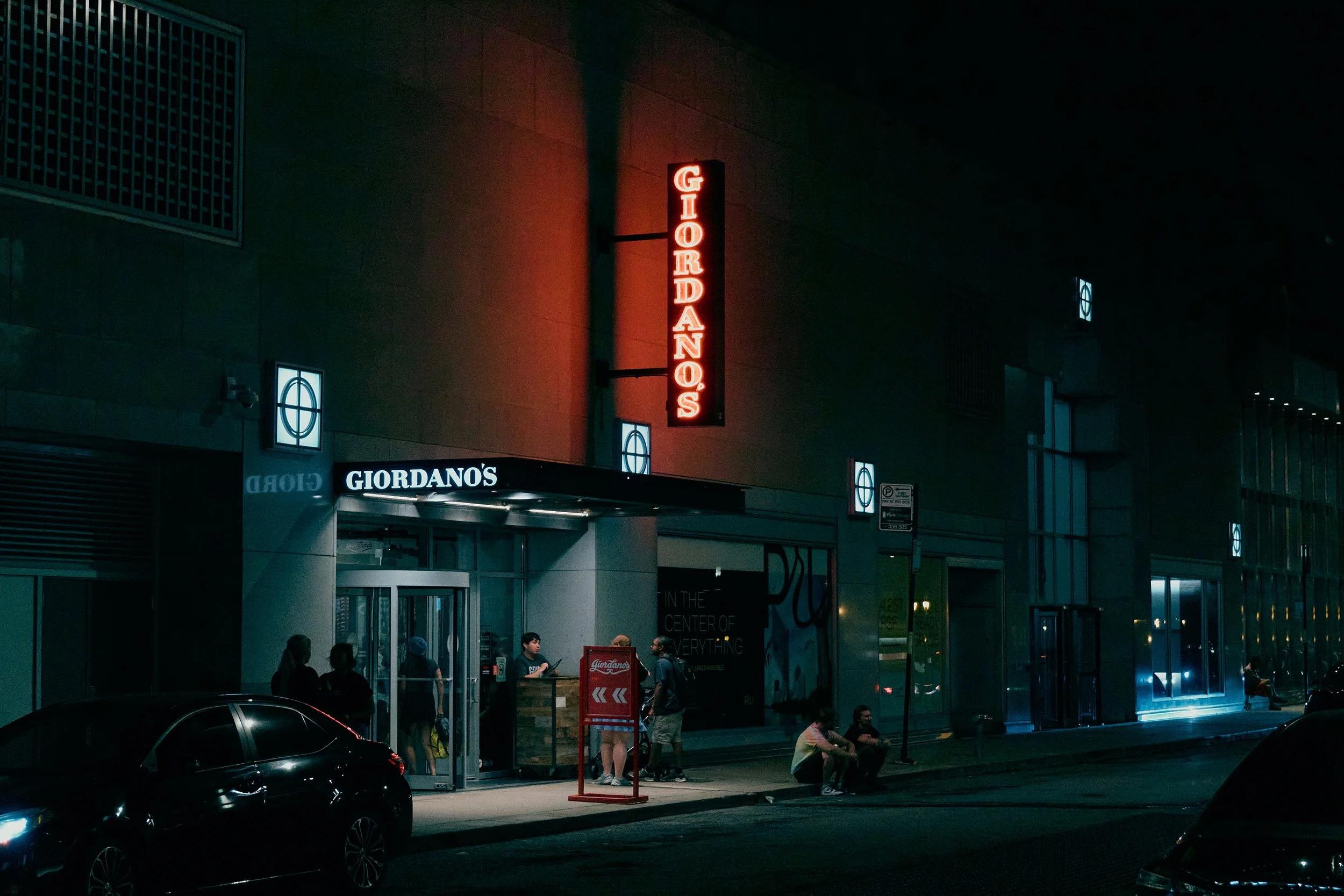 Night view of Giordano's restaurant with illuminated vertical and horizontal signs, a few people sitting and standing outside, and cars parked along the street.