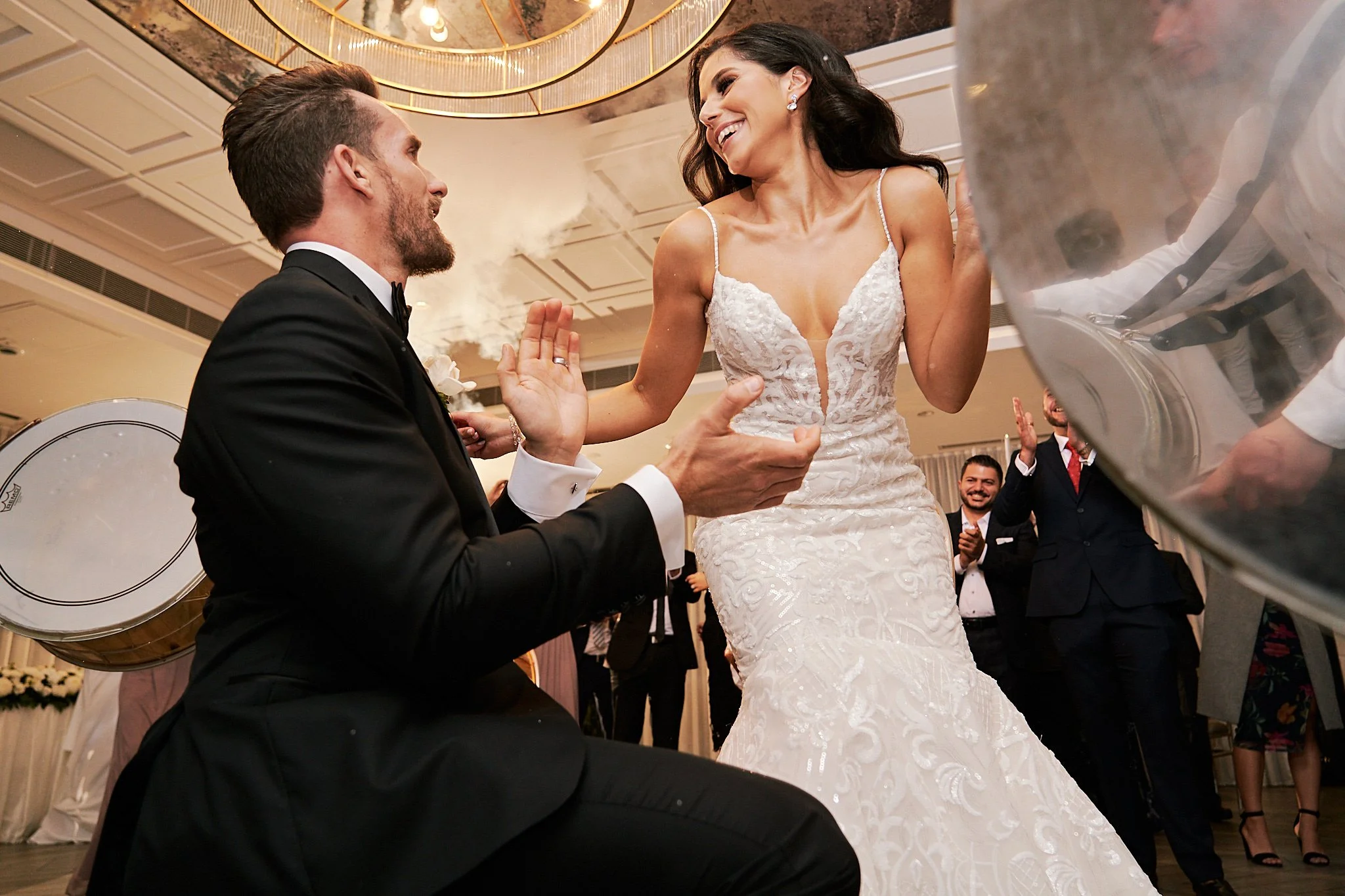 A bride and groom dancing at a wedding reception. The groom is kneeling and wearing a black tuxedo, while the bride is standing and wearing a white lace wedding gown. Guests in suits and dresses are clapping and smiling in the background.