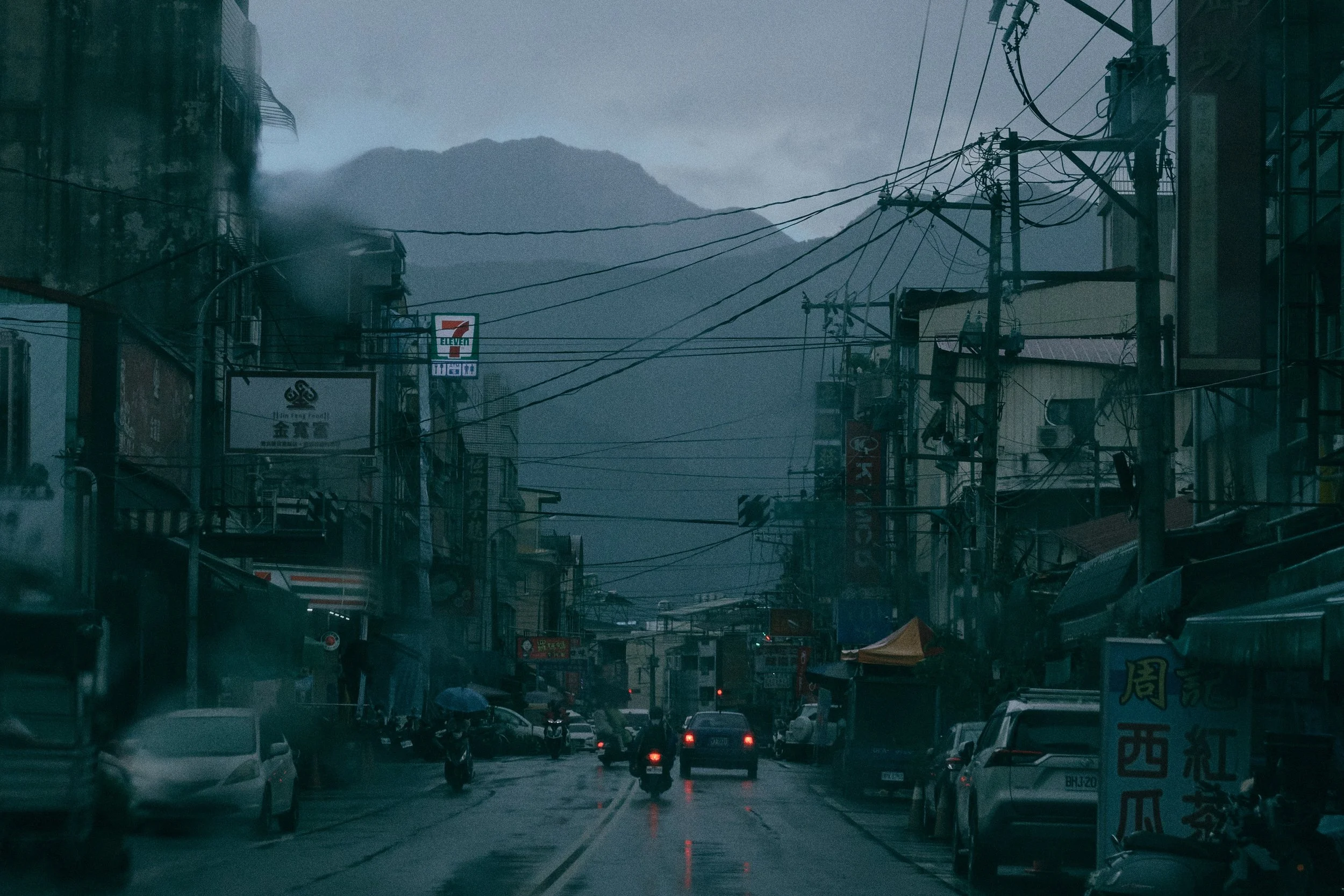 A rainy, urban street scene with cars, motorcycles, signs, storefronts, overcast sky, power lines, and mountains in the background.