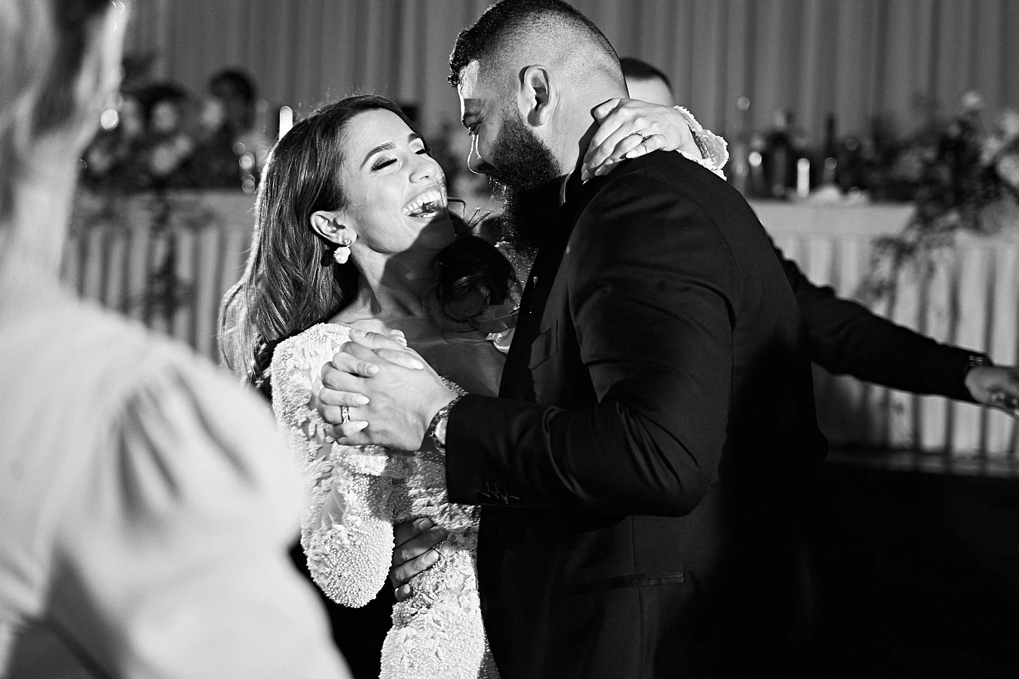 A man and woman are dancing and laughing happily at what appears to be a wedding reception, with other people and decorations in the background.