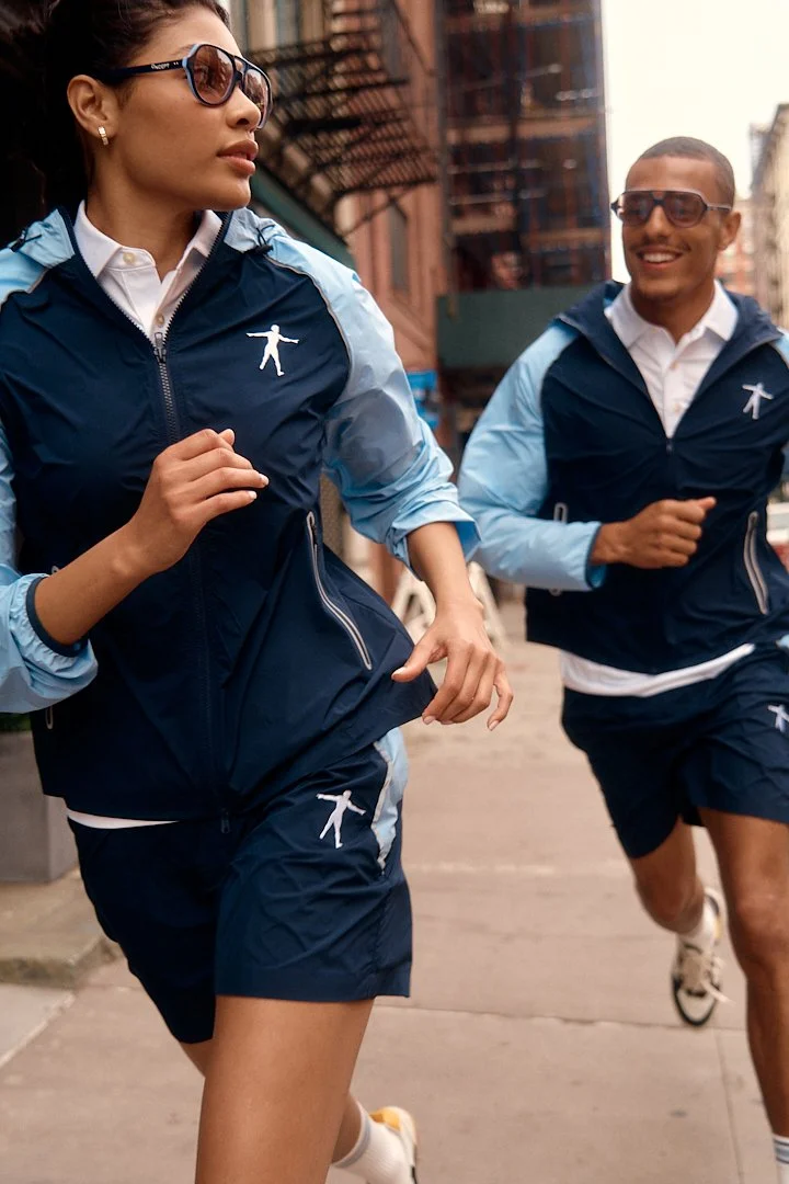 A man and woman running outdoors on city streets, both wearing matching navy and light blue athletic jackets, shorts, and sunglasses.