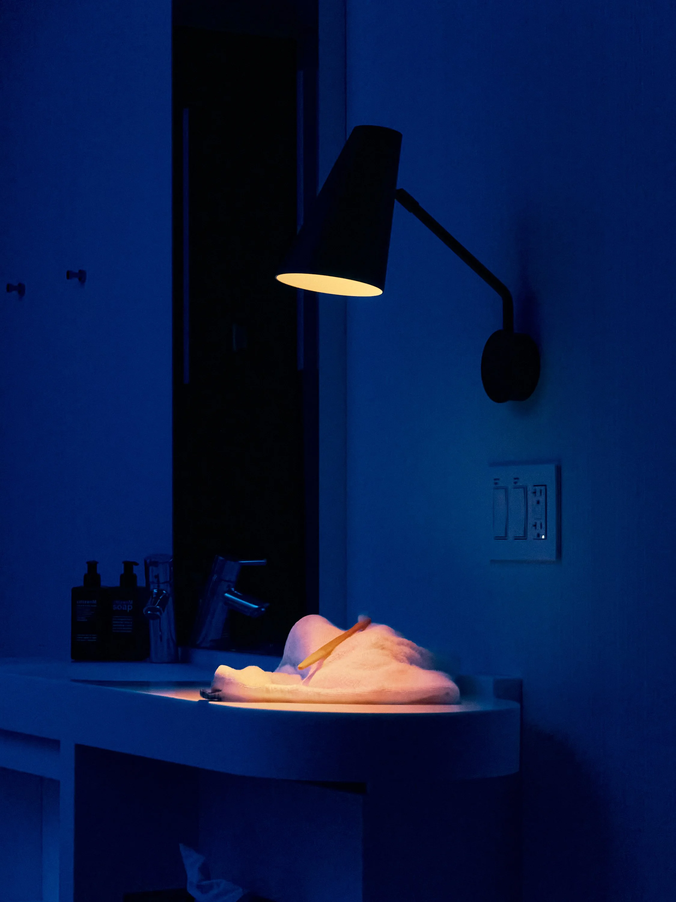 A dimly lit bathroom with a wall-mounted lamp illuminating a towel and a toothbrush on the counter, and a dark open door reflected in the mirror.