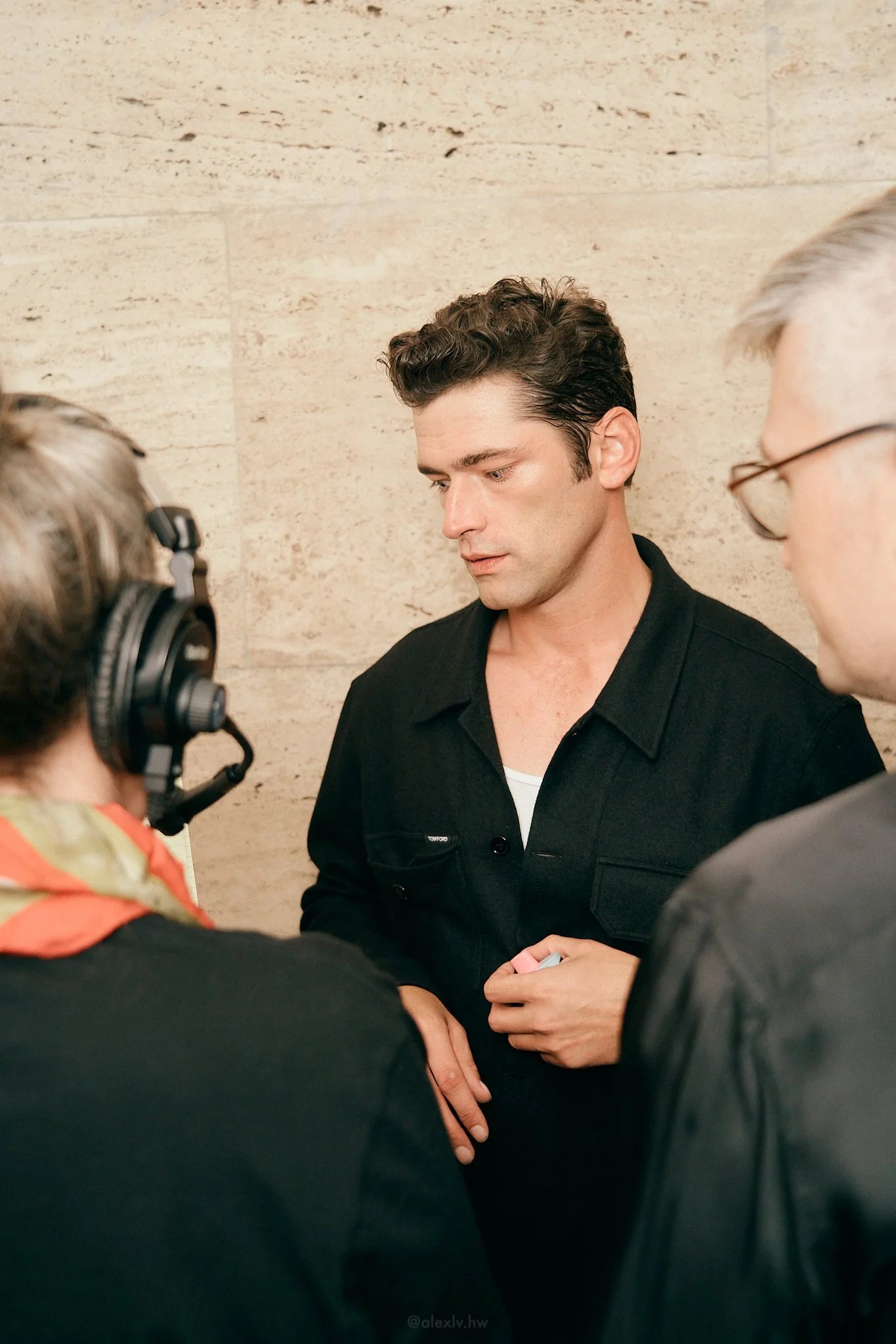 A man with dark hair and a black jacket being interviewed by two people, one with headphones, against a beige stone wall.