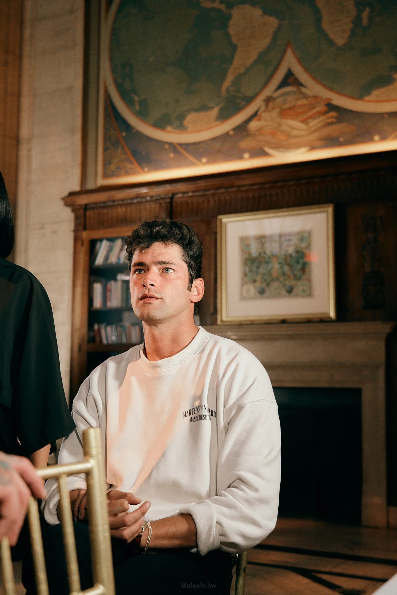 A young man with dark hair sitting in a room with wooden paneling, framed artwork, and a large map on the wall behind him. He is wearing a white sweatshirt with text on it and has his hands clasped in his lap.