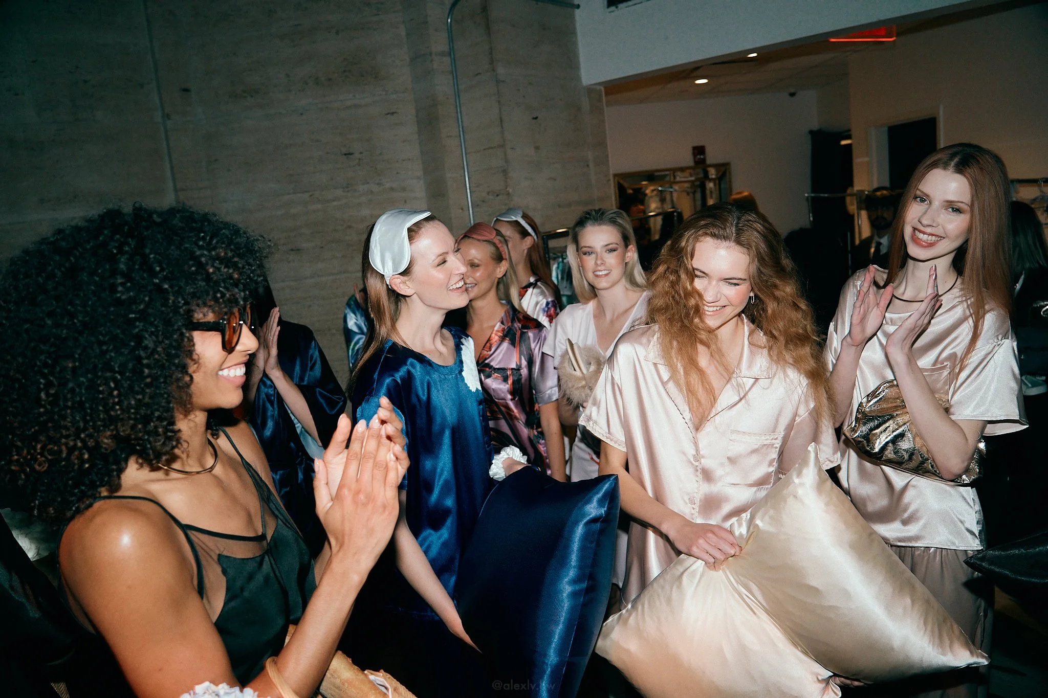Group of women in silk pajamas and robes laughing and chatting at a sleepover or pajama party, some holding pillows.