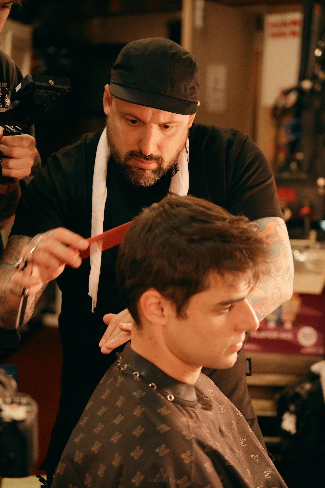A barber cutting a young man's hair in a barbershop. The barber is wearing a black shirt, a black cap, and has tattoos on his arms. The young man is seated and wearing a barber cape, looking down.