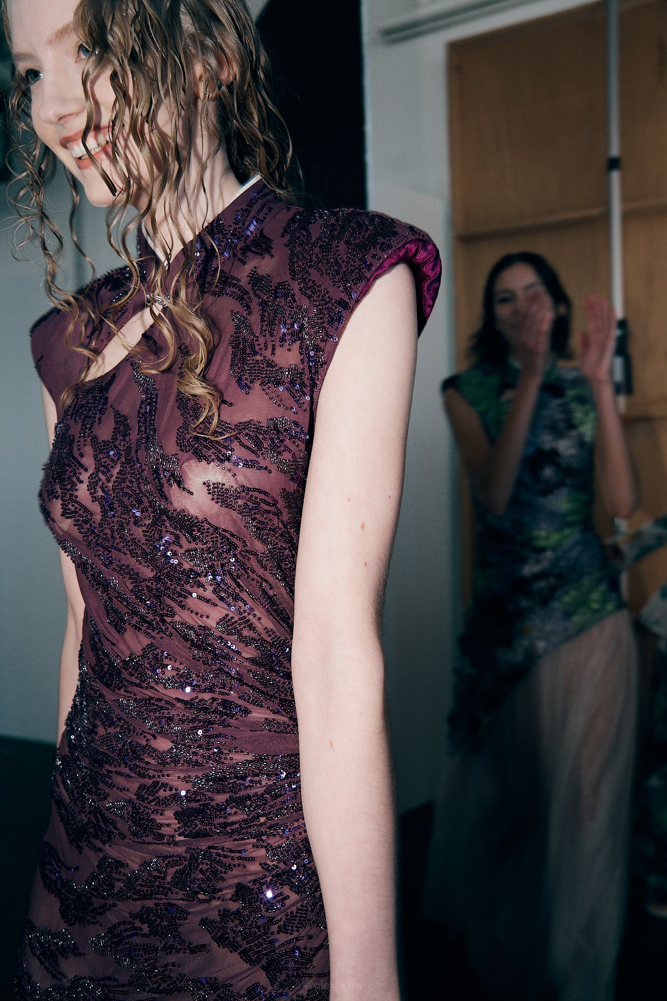 A woman in a sheer, purple, sequined dress with ruffle sleeves, smiling, with another woman clapping and smiling in the background.