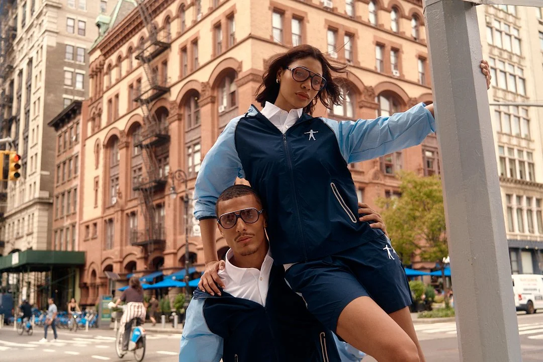 A young woman and man wearing matching blue and white athletic outfits with sunglasses, posing on a city street with historic buildings and pedestrians in the background.