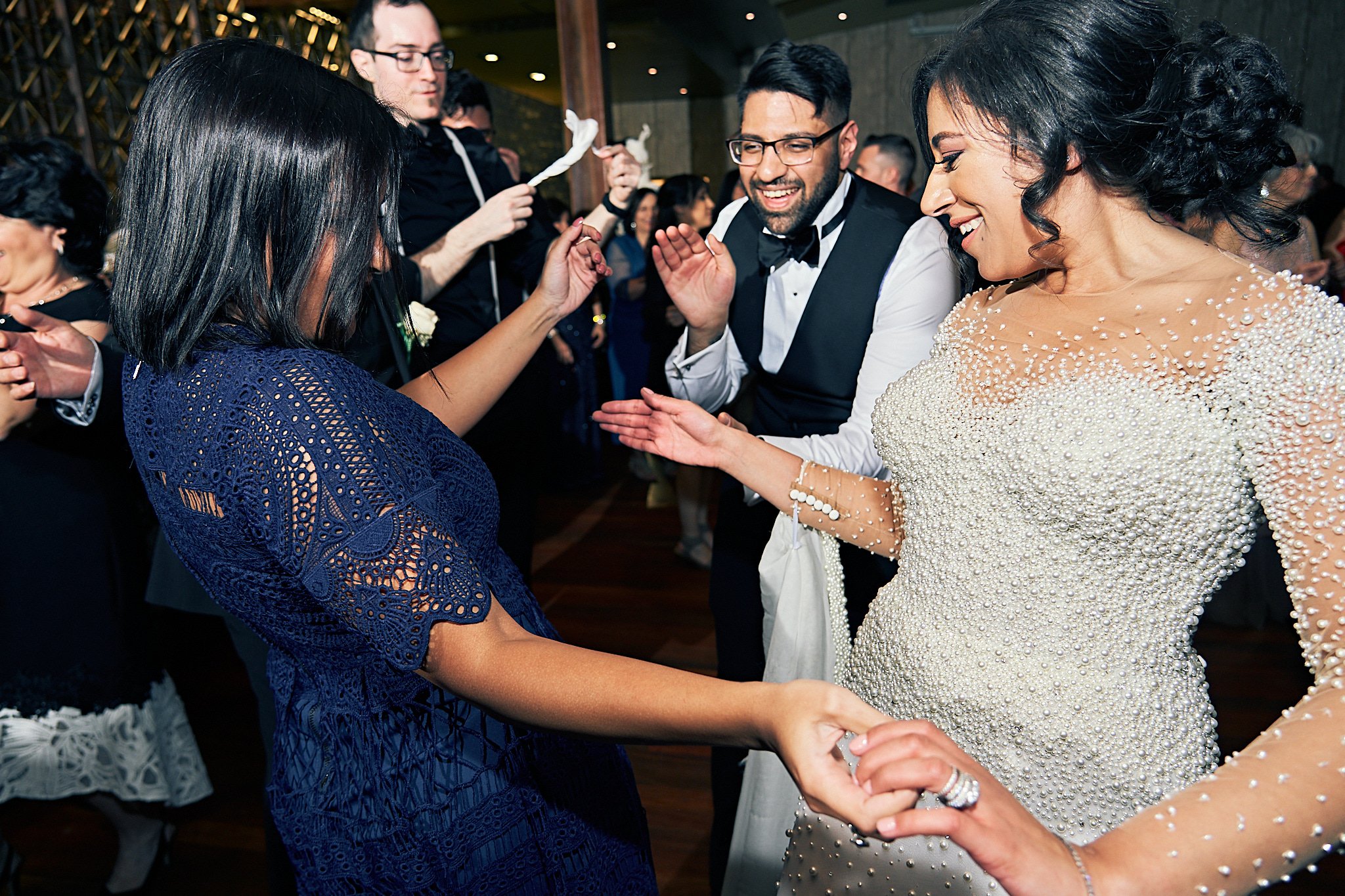 People dancing and celebrating at a wedding reception with a bride in a pearl-embellished gown and a groom in a tuxedo.