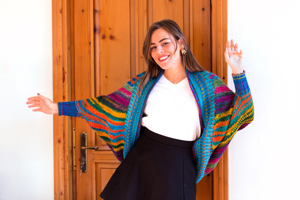 a young white woman with dark blonde hair frolics in front of a wooden door. She's wearing a white tshirt and black skirt and a colorful cocoon jacket with multiple colors - fuschia, purple, aqua, royal blue, lime green, dark green, and orange stripes of varying widths, with a herringbone texture