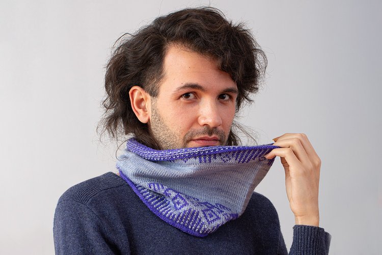 An olive-skinned man with medium-length dark curly hair and dark stubble holds up the edge of his Blue-and-grey colorwork cowl. He has a smoldering look in his eye