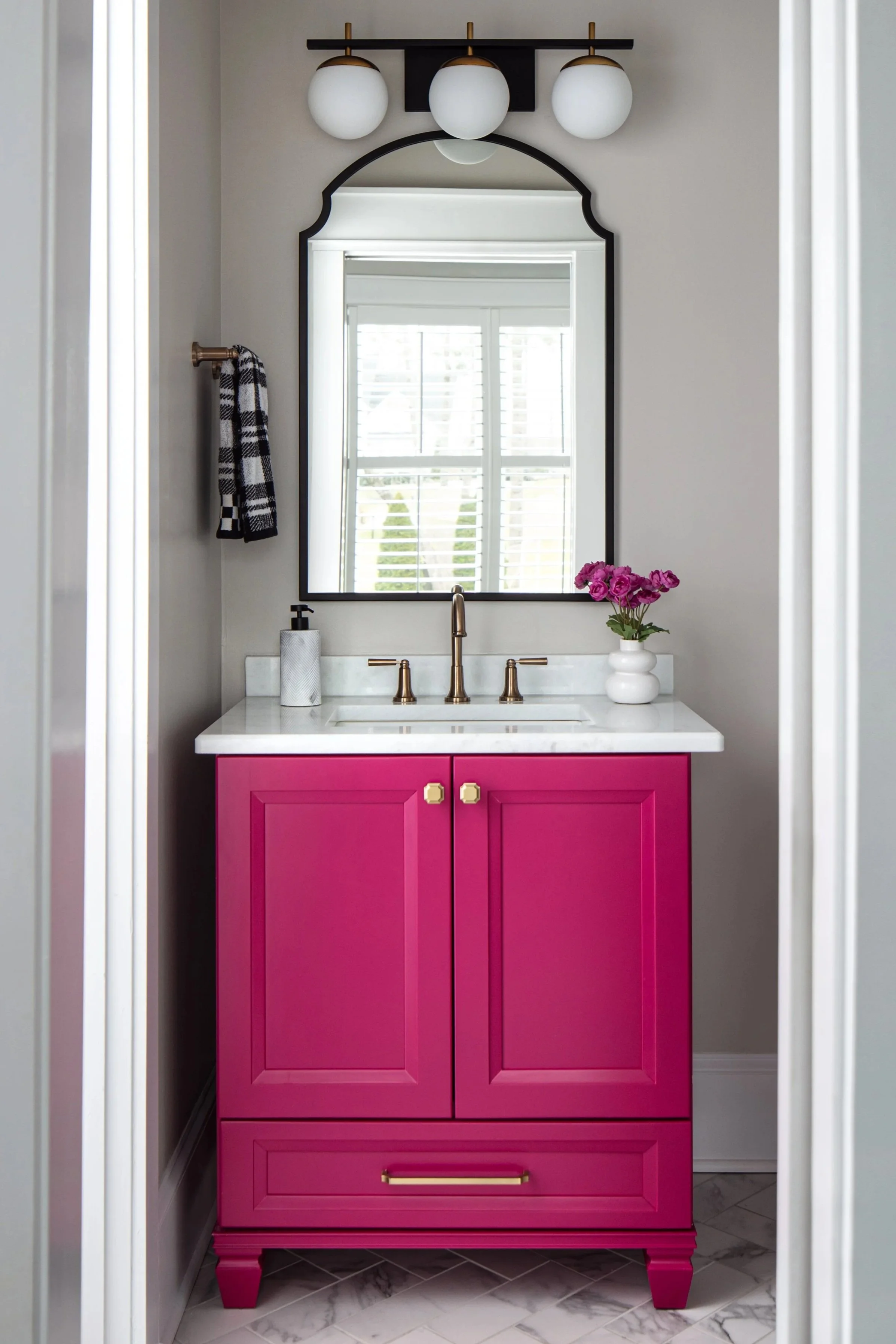 A bathroom renovation by Cathy Green Interiors feature a pink vanity, an arched mirror, and a fun light fixture.