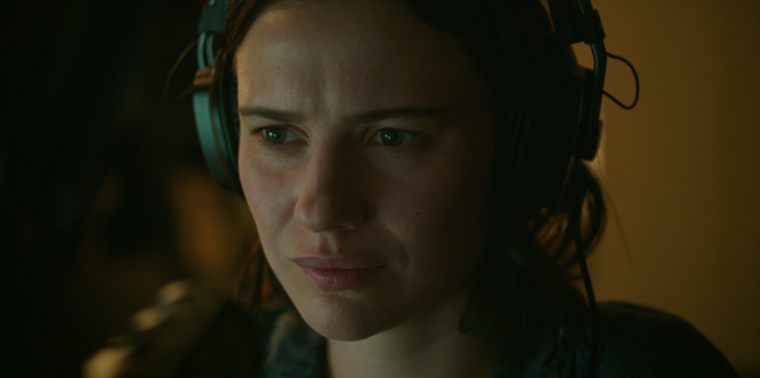 Close-up of Lily Sullivan in headphones in Monolith, warm low-key lighting as the story darkens.