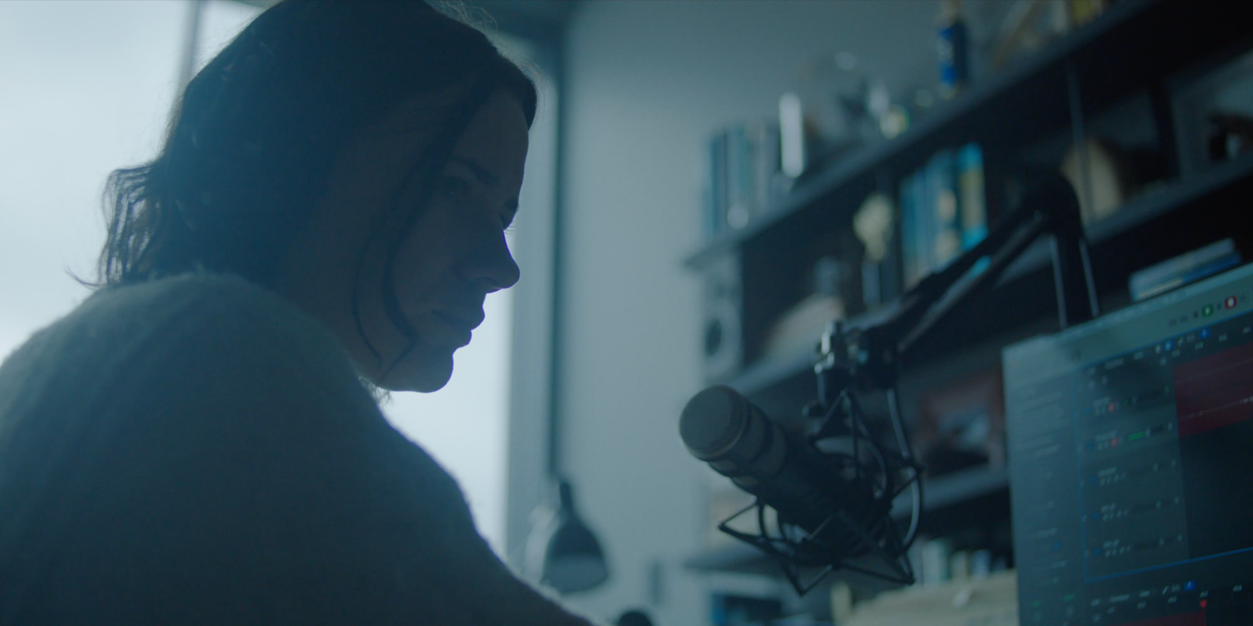 "Over-the-shoulder frame from Monolith of Lily Sullivan at the microphone, shot in cool available window light."