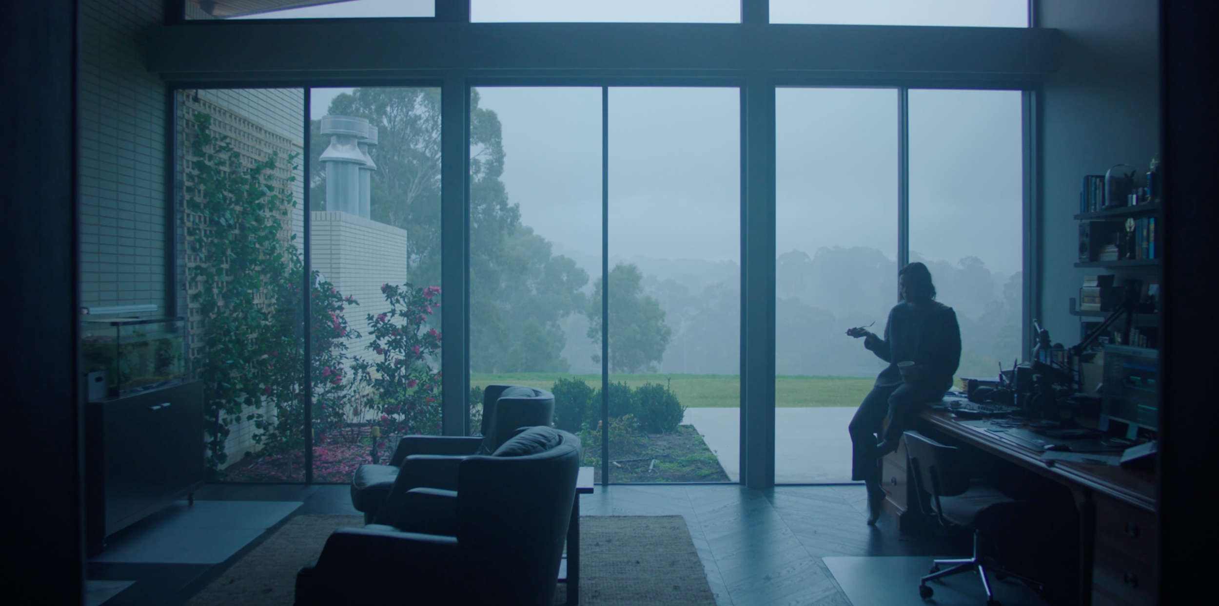 Wide interior frame from Monolith — the character alone against fog and rain outside the Adelaide Hills house's glass walls.