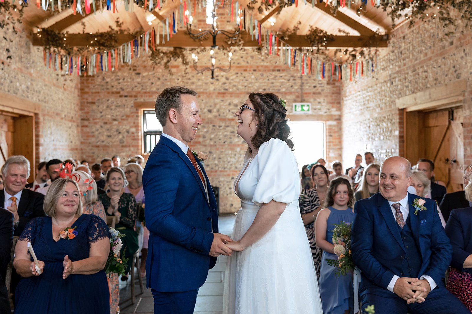 Bride and groom laughing as they say their vows in front of guests in a rustic barn wedding venue