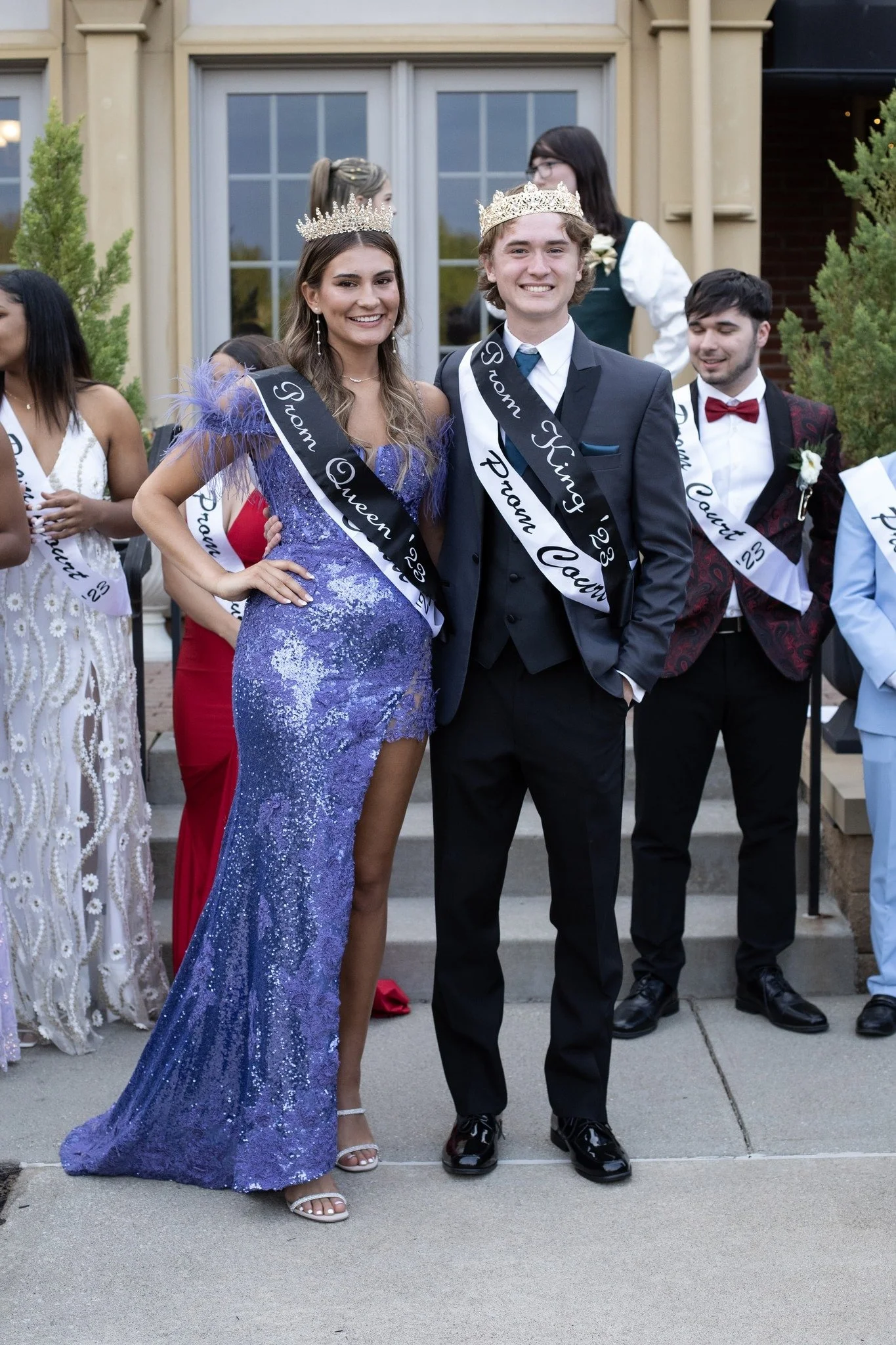 A group of young adults at a prom celebration, with two wearing sashes that say 'Prom Queen 23' and 'Prom King 23' and crowns. The woman is in a purple sequin gown, and the man is in a black tuxedo. They are standing on a sidewalk in front of a building with large windows and greenery.