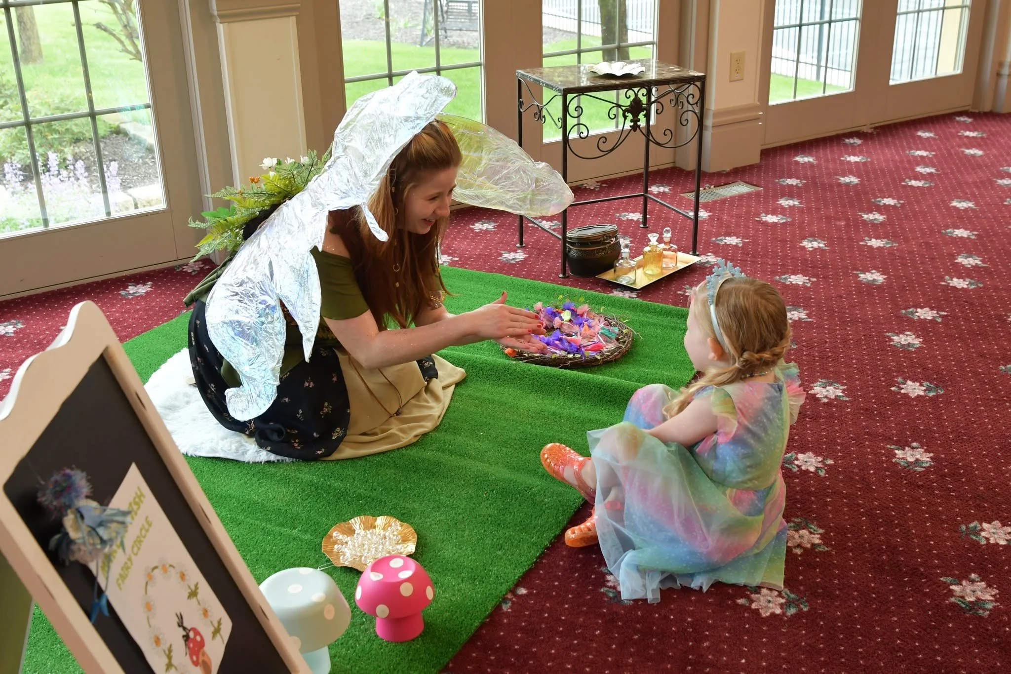 A fairy-tale themed room with large glass doors, a woman dressed as a fairy with aluminum foil wings and a flower crown, and a young girl in a rainbow dress and fairy wings sitting on a green carpet, participating in a craft activity with flowers.