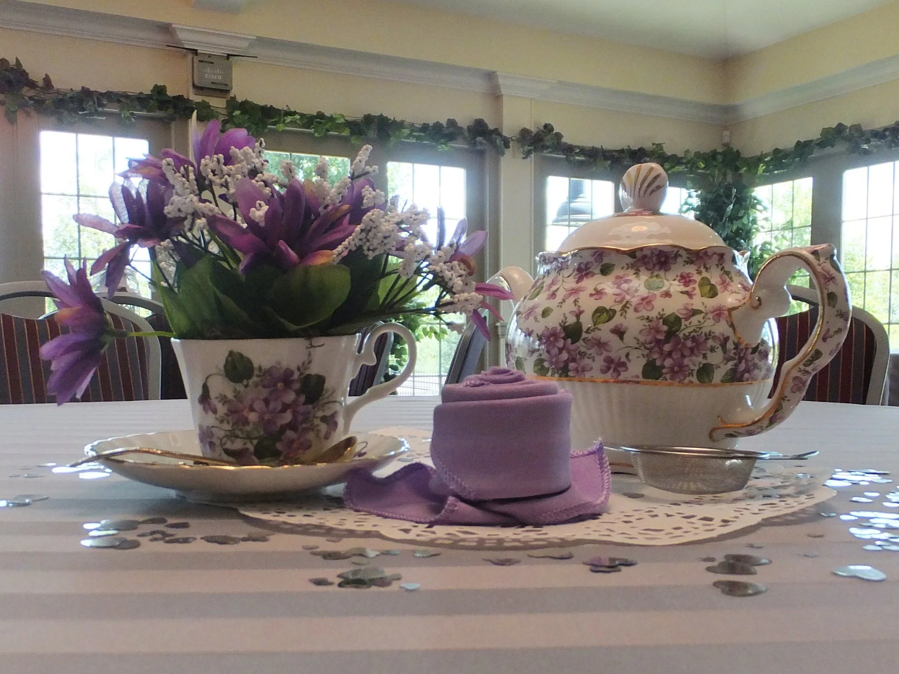 A dining table set with a floral teapot, a matching floral cup, a vase with purple and white flowers, a purple napkin, and scattered confetti, with a backdrop of large windows and greenery outside.