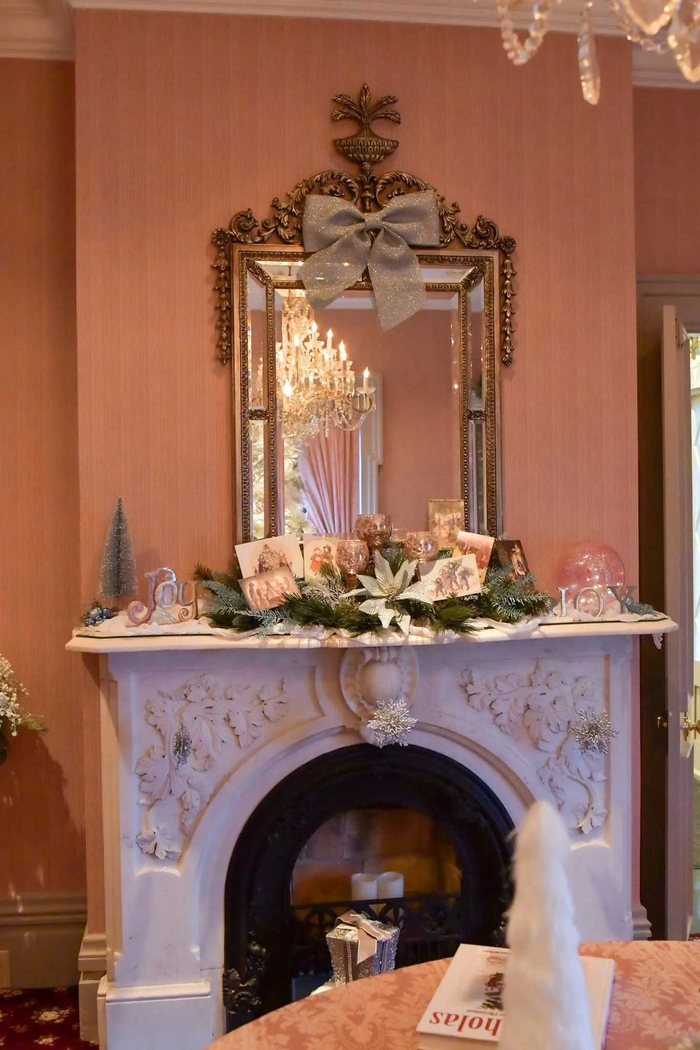 A decorated fireplace mantel with holiday decorations including a large silver bow, Christmas cards, ornaments, and a small gift box, all set against a pink wall with a mirror and chandelier.