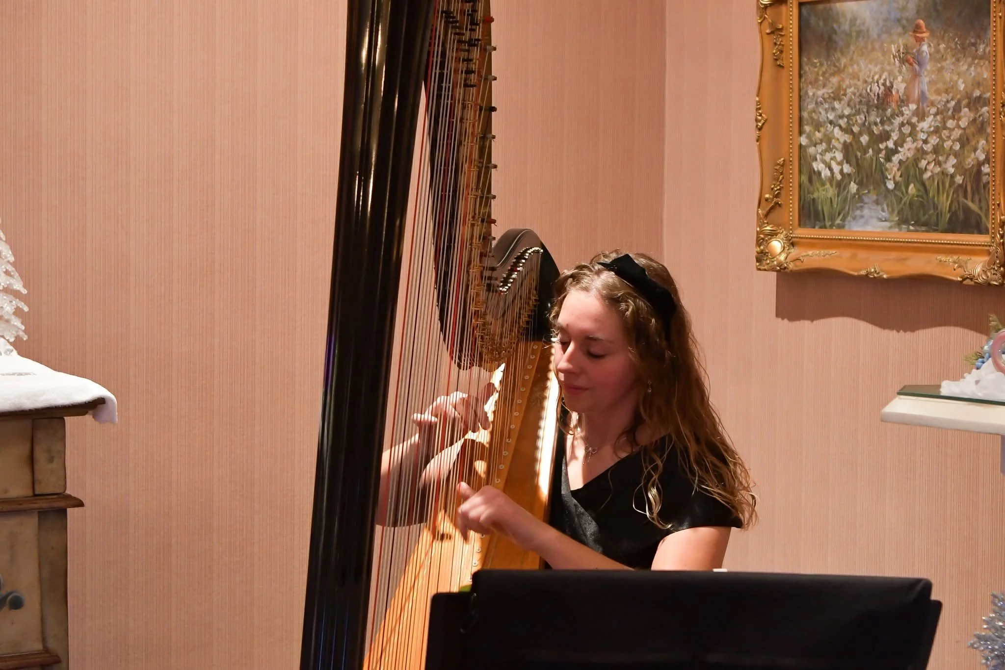 A young woman with long wavy hair playing a harp indoors, with a framed painting of a woman in a white dress and hat in a field of flowers hanging on the pink wall behind her.