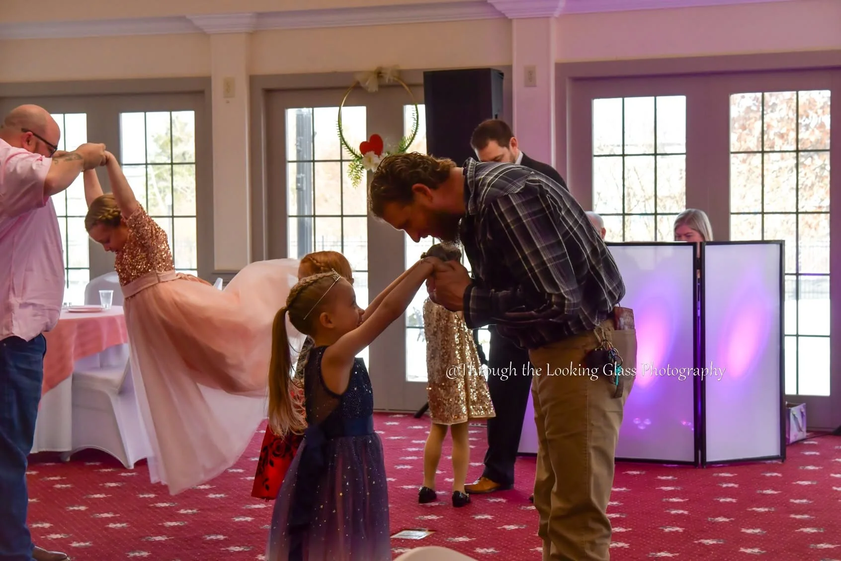 A man is giving a kiss on the forehead to a young girl at a social event, with other children and adults dancing and enjoying themselves in the background.