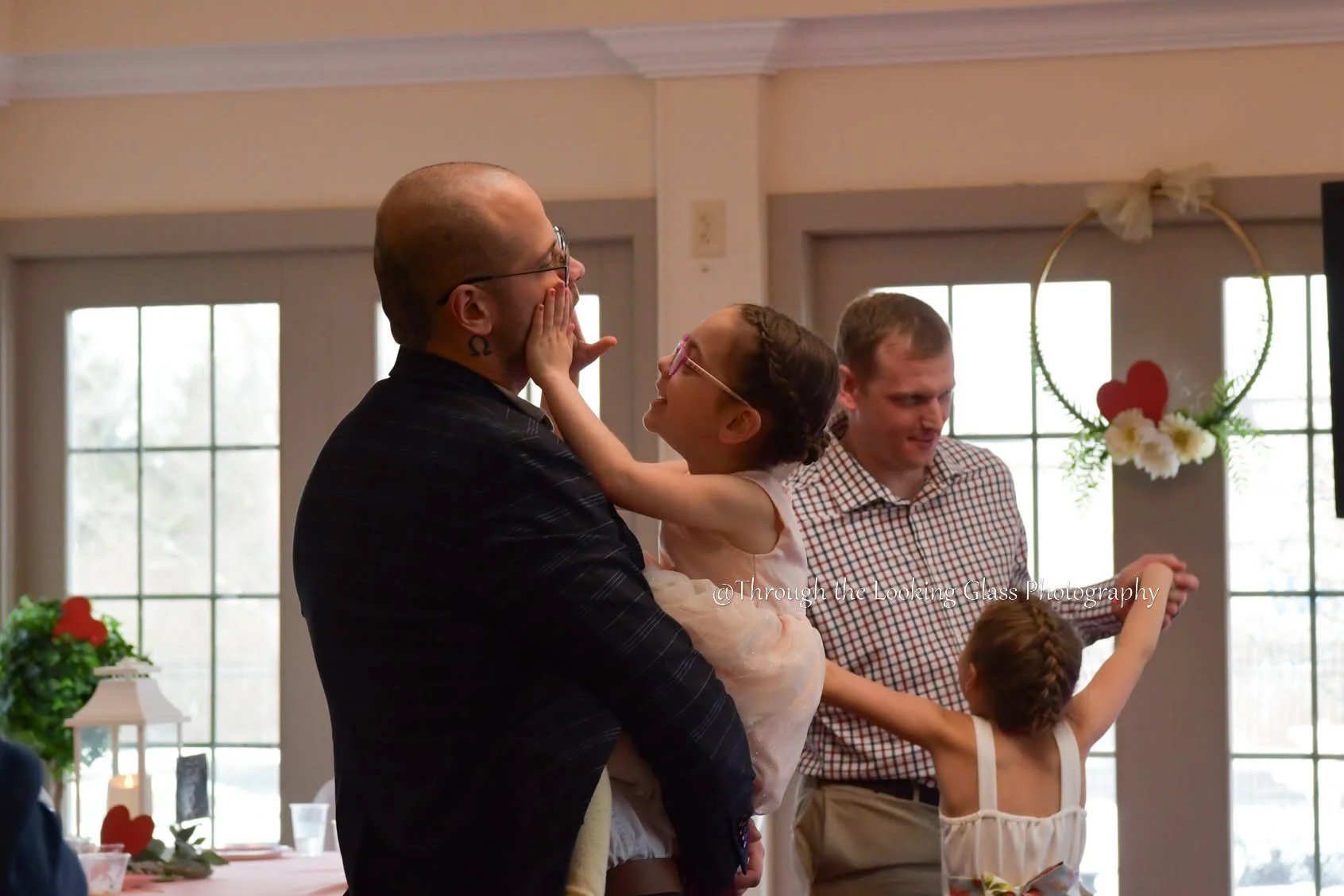 A man holding a young girl with glasses, who is touching his face, while smiling and looking at him. In the background, a man in a checkered shirt is standing near a young girl with braids, inside a room decorated with flowers and heart-shaped decorations on the windows.