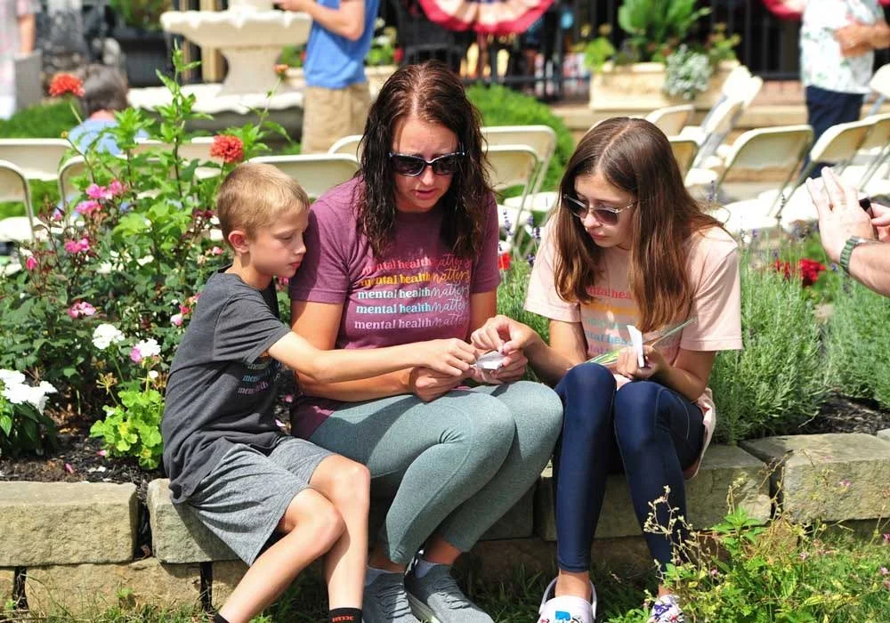 A woman, a boy, and a girl sitting on a stone bench in a garden with flowers, looking at a smartphone and a note, with people and outdoor chairs in the background.