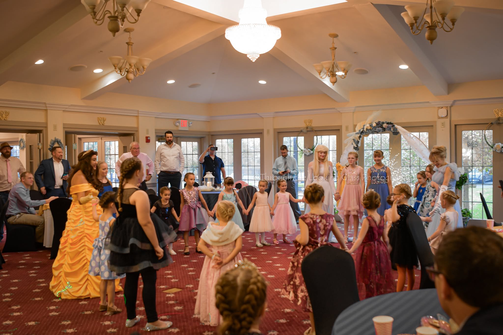 A room filled with children and adults during a celebration, with children dressed in fancy dresses and adults watching or taking photos. There is a decorated arch and a chandelier hanging from the ceiling.