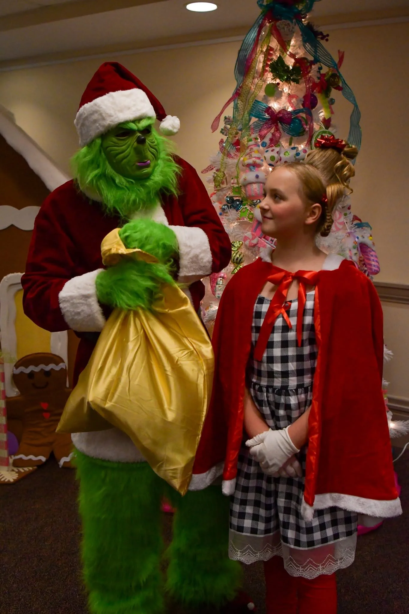 A person dressed as the Grinch wearing a Santa hat and green fur, talking to a young girl dressed as Cindy Lou Who in a red cape and checkered pajamas, celebrating Christmas with a decorated tree in the background.