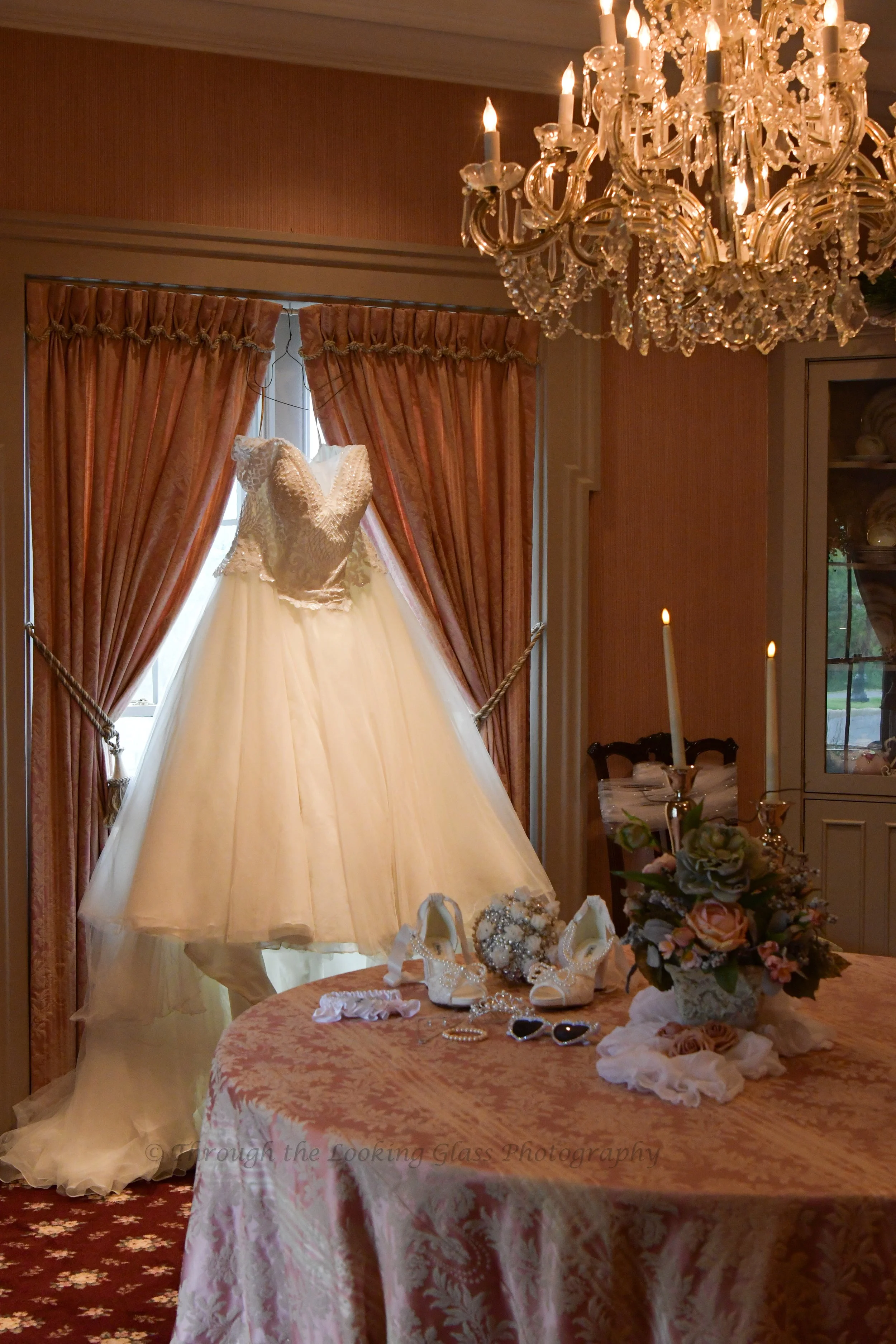 A bridal dress displayed on a mannequin in front of a window with pink curtains. A chandelier hangs from the ceiling. A table covered with a pink floral tablecloth holds a bouquet of flowers and various accessories, including jewelry, shoes, and sunglasses.
