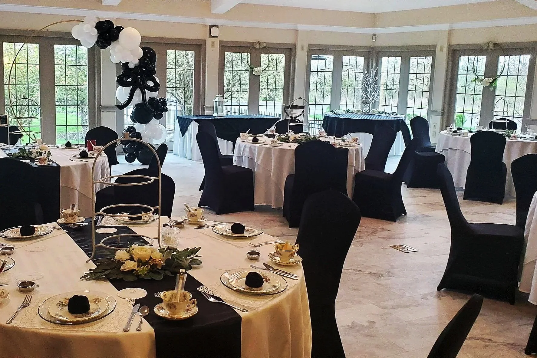 Decorated banquet hall with round tables covered in white and black tablecloths, set for a meal, with black chairs, floral centerpieces, and balloon arrangements, large windows with natural light, and elegant wall accents.