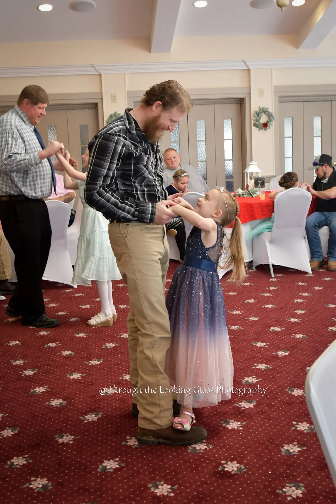 A man with a beard and a young girl in a princess dress are dancing at a celebration in a decorated hall. Other people are sitting or dancing in the background.