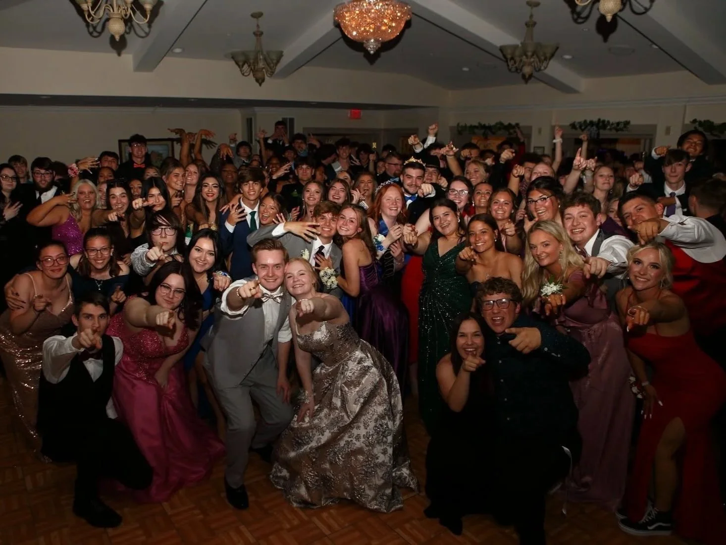 A large group of young people in formal attire at a celebration, all posing with fists pointed forward, in a decorated ballroom with chandeliers.