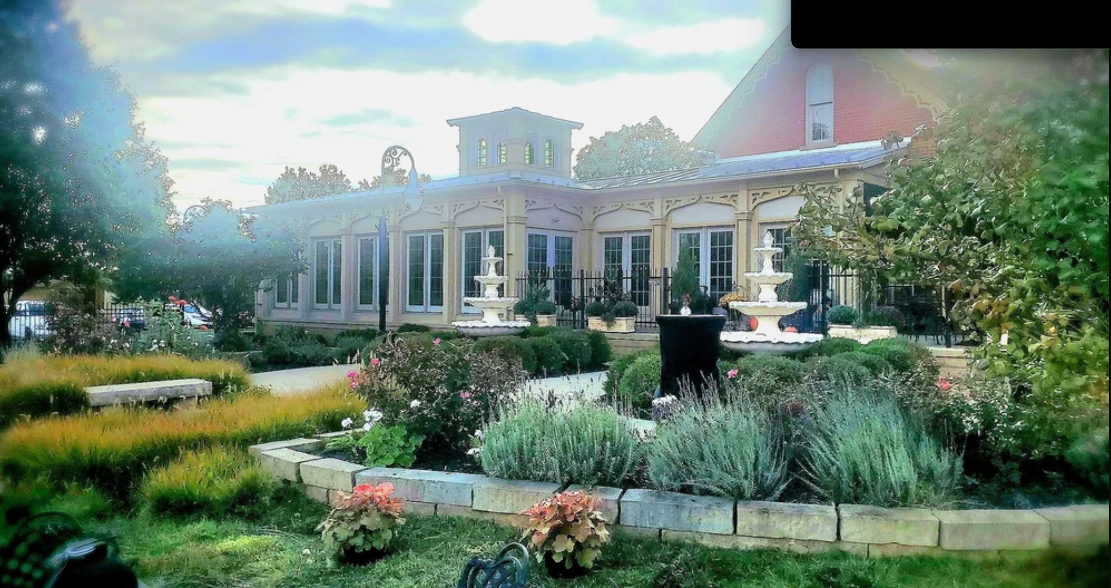 A charming house with large windows, a garden with landscaped plants, trees, and a stone path. A fountain is visible in the lawn area, and there's a wrought iron fence around the property.