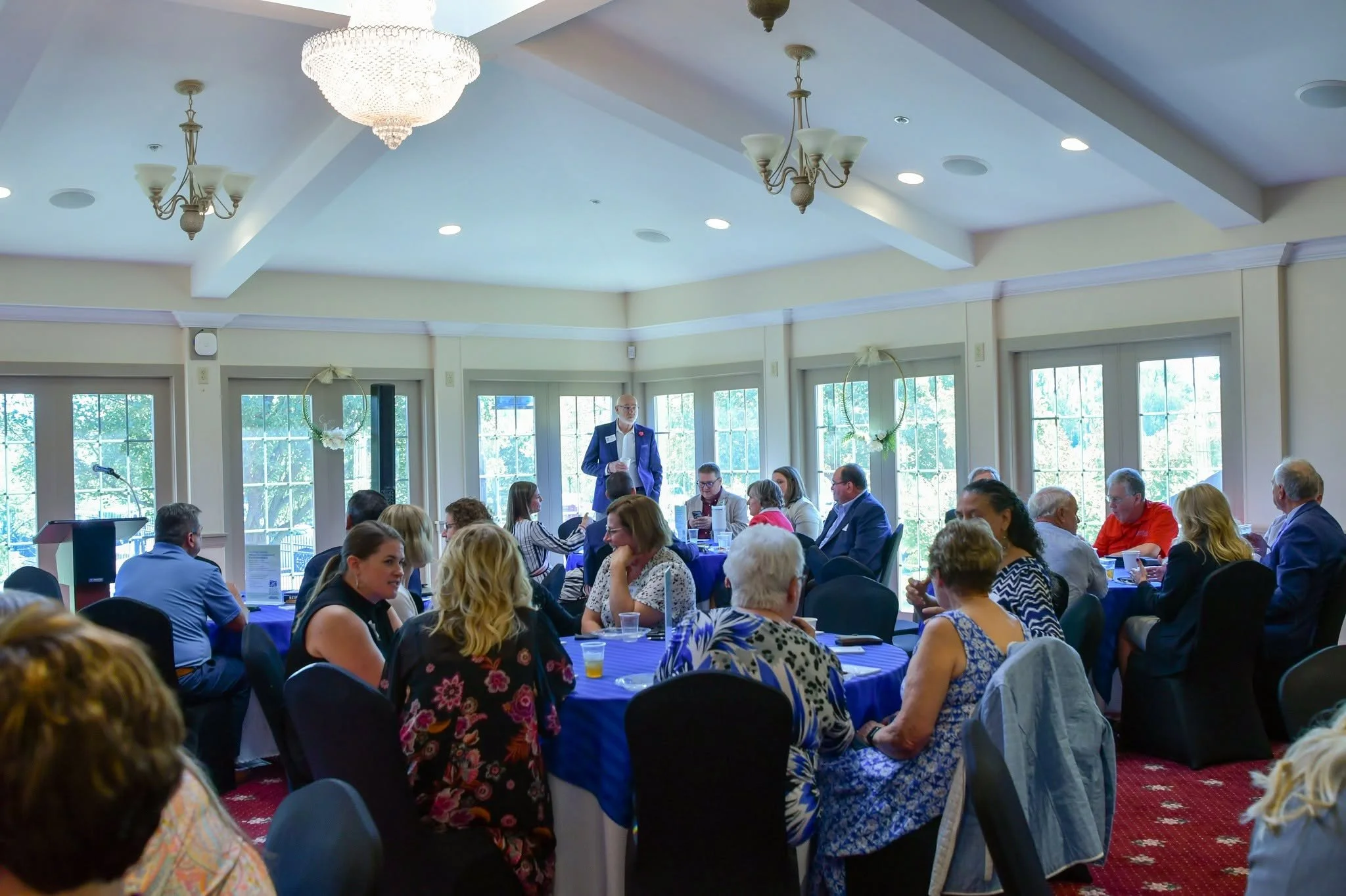 A group of people seated at round tables in a banquet hall, attending a formal event or conference, with a man standing and speaking at the front. The room has large windows, chandeliers, and decorated wreaths hanging on the walls.