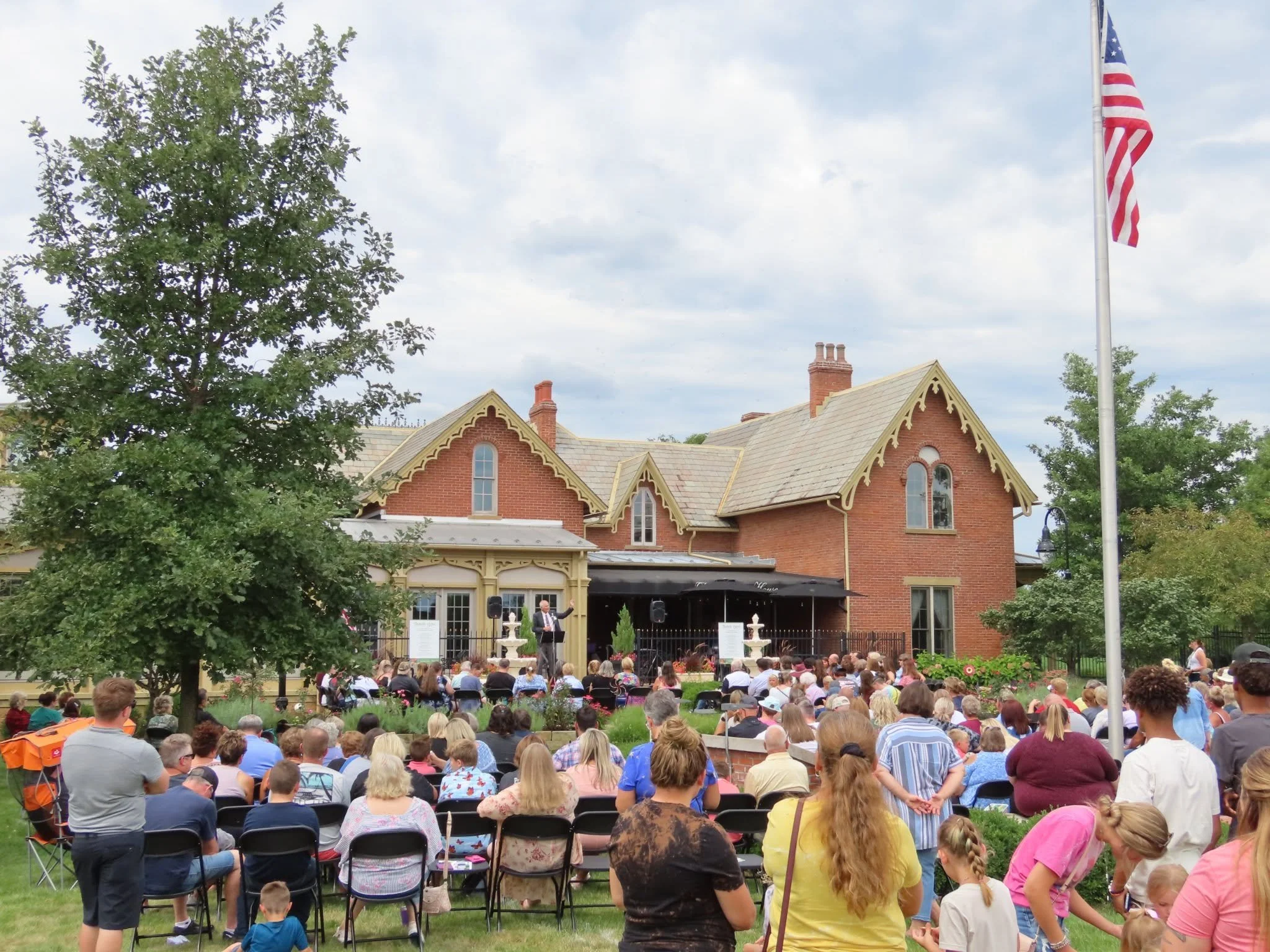 A large crowd of people seated outdoors in front of a historic brick building with pointed roof accents, while a man stands speaking on a small stage. An American flag on a tall pole is visible on the right side of the image, and a large green tree partially obscures the building. It appears to be a community or public event, possibly a concert or speech.