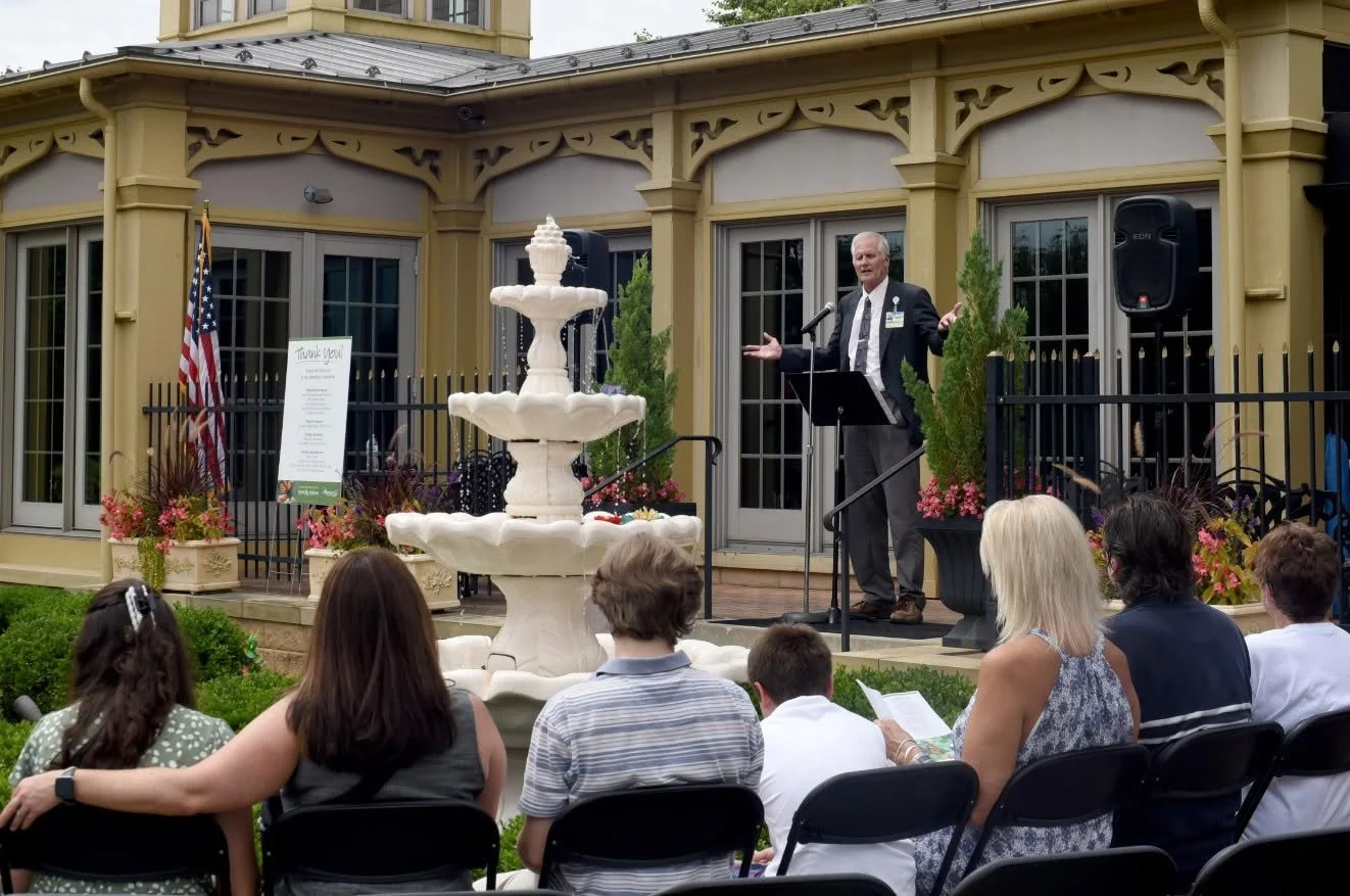 A man in a suit giving a speech or presentation outdoors on a small stage, with an audience seated in front. The setting includes a decorative fountain, potted plants, and an American flag. The event is taking place in front of a yellow building with large windows.