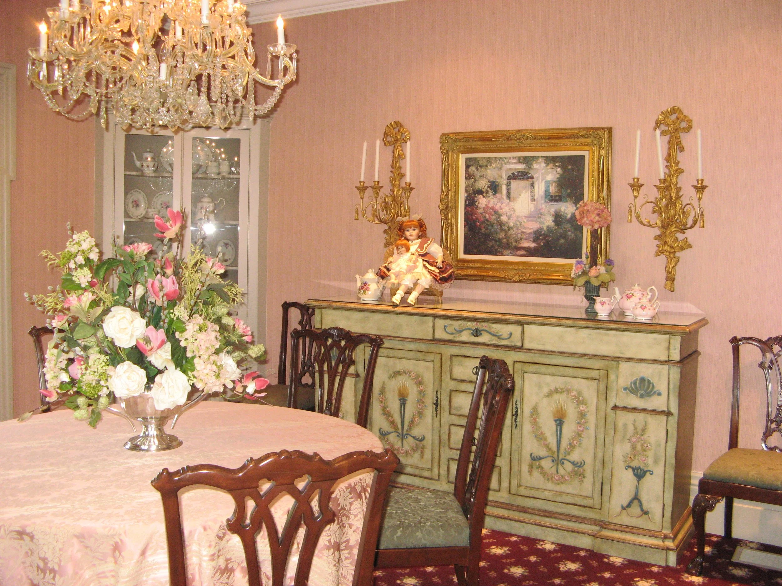A dining room with a large floral centerpiece, a crystal chandelier, an ornate green-painted sideboard decorated with floral motifs, a painting of a garden scene, porcelain tea sets, and a doll seated on the sideboard.