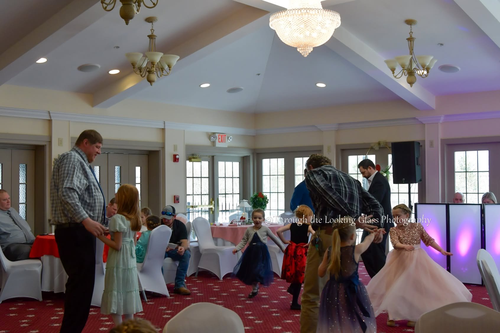 Children and adults dancing and socializing in a decorated banquet hall with chandeliers, tables, and chairs.