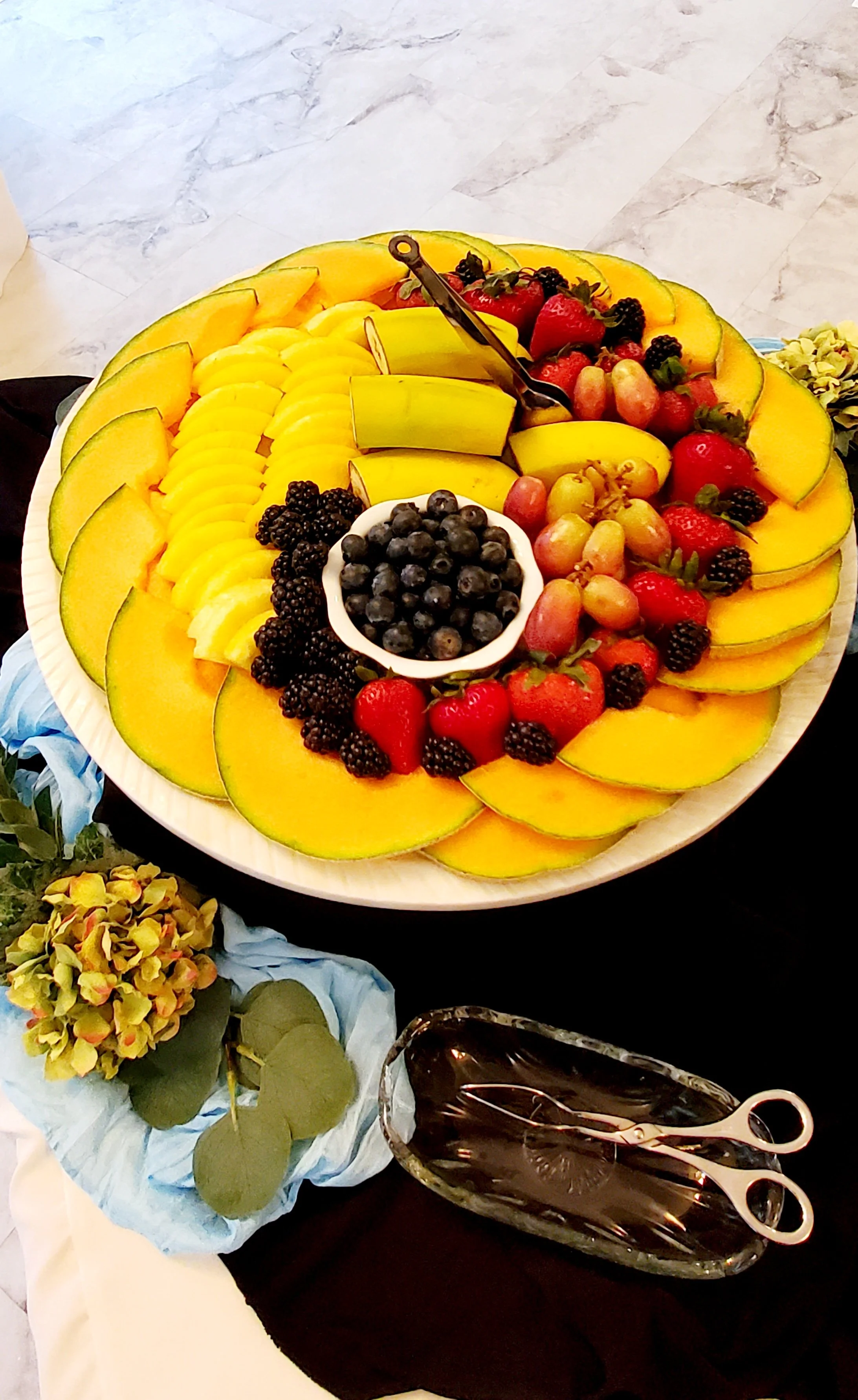 A large round platter of sliced cantaloupe, strawberries, blueberries, blackberries, and watermelon, garnished with fresh green herbs, with a bowl of blueberries in the center. There is a serving spoon on the platter. Nearby, on a black tablecloth, is a glass dish with chocolate sauce and a pair of small scissors with white handles, along with a small pot of hydrangea flowers and a sprig of leaves.