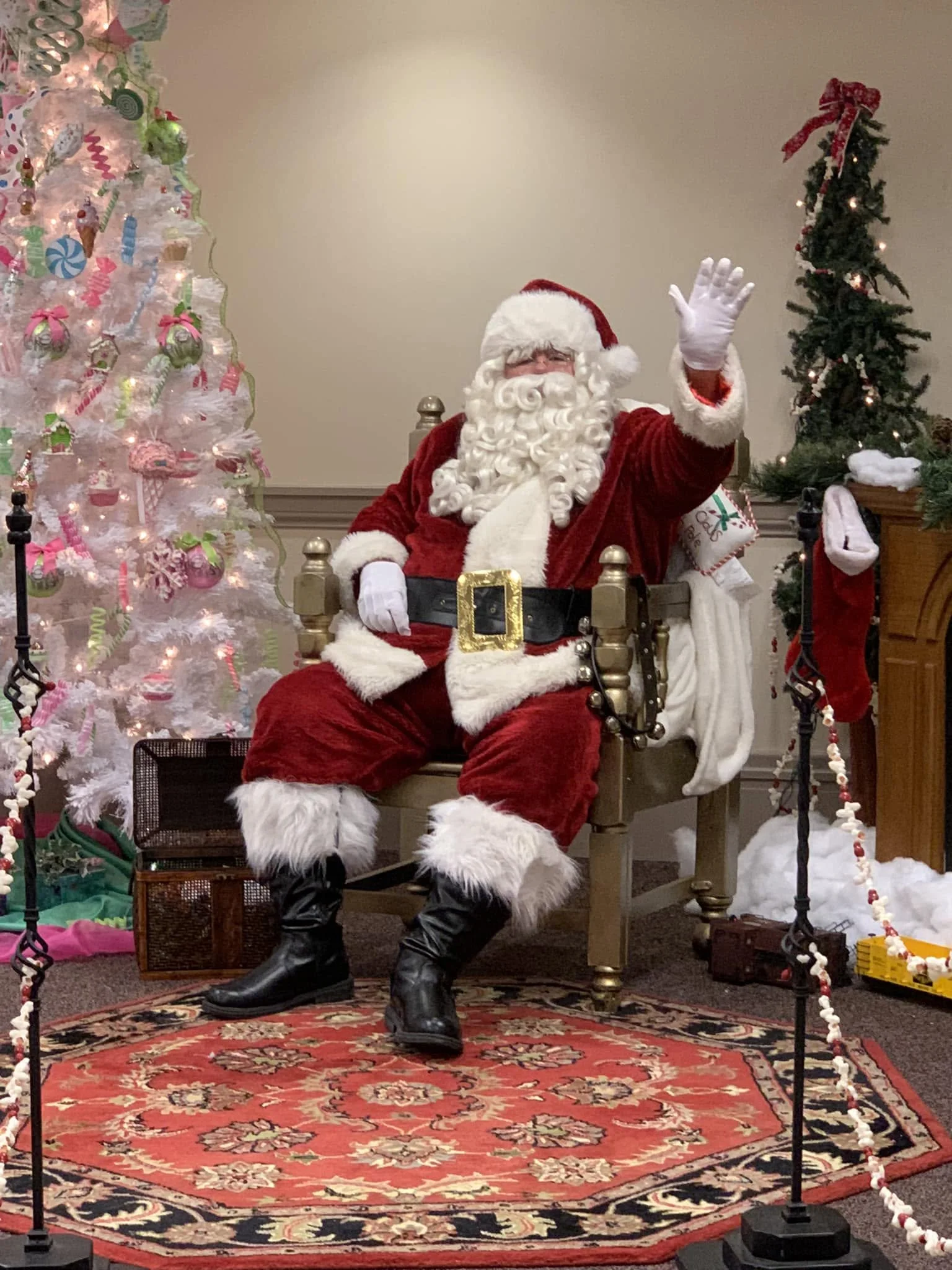Santa Claus with a white beard, red suit, and black boots sitting on a chair, waving. Behind him are decorated Christmas trees, one pink with colorful ornaments and lights, and another green with a red ribbon on top and lights. Santa is gesturing to the camera. The scene is set on a red patterned rug, with a red and white rope barrier in front.