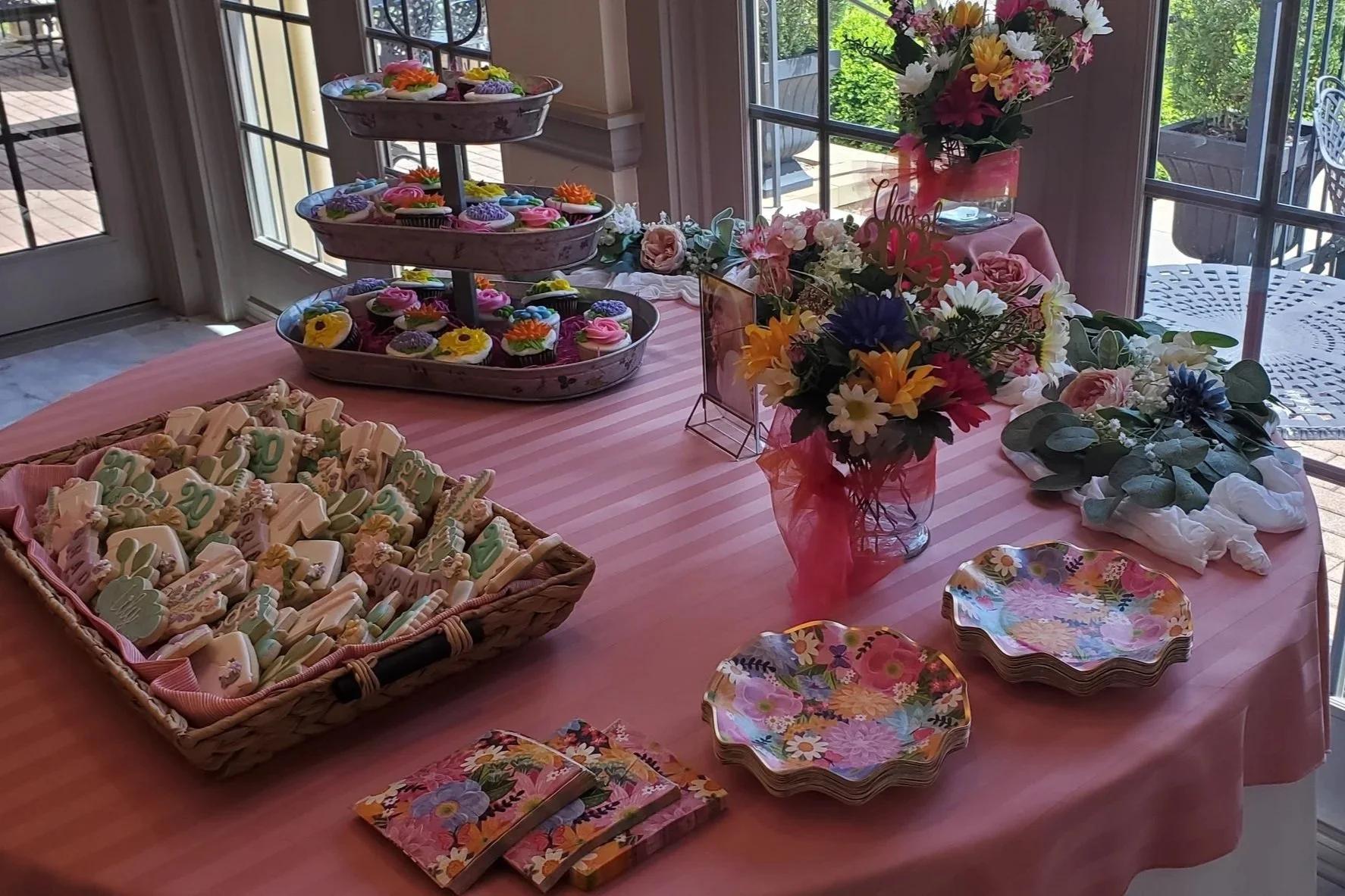 A table decorated with a pink tablecloth, floral arrangements, plates, sundaes with colorful frosting and sprinkles, and small wrapped candies. It appears to be set for a celebration, possibly a birthday party.