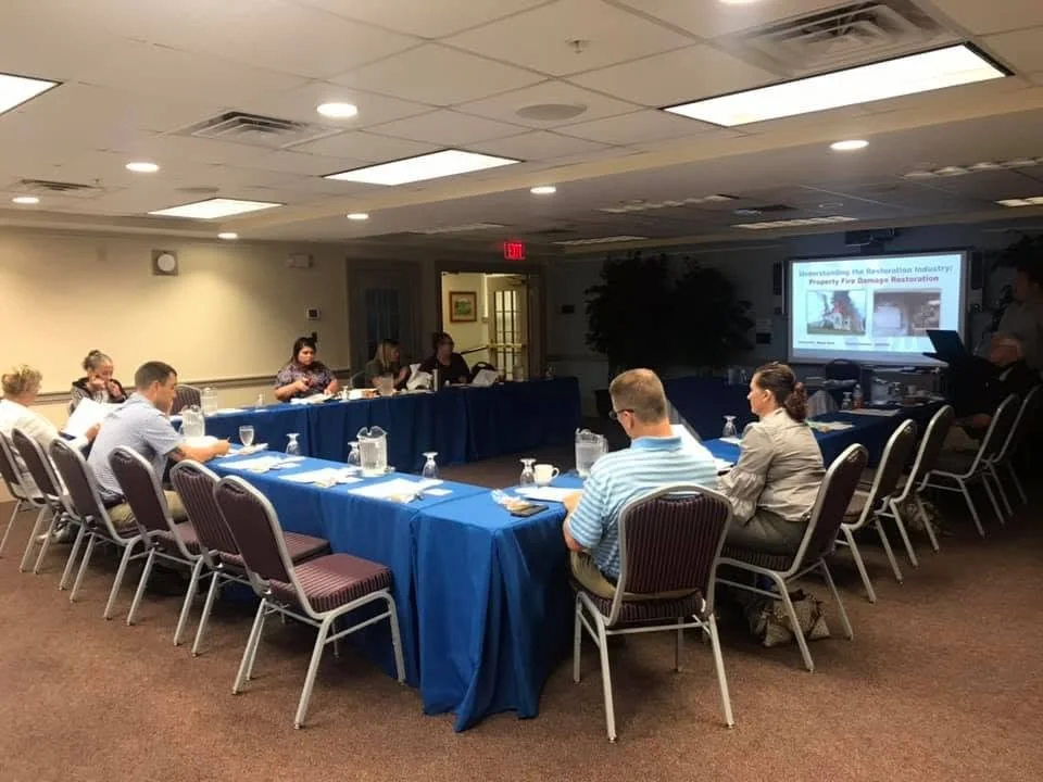 A group of people attending a meeting in a conference room, seated at U-shaped tables with blue tablecloths, with presentation slides projected on a screen at the front.