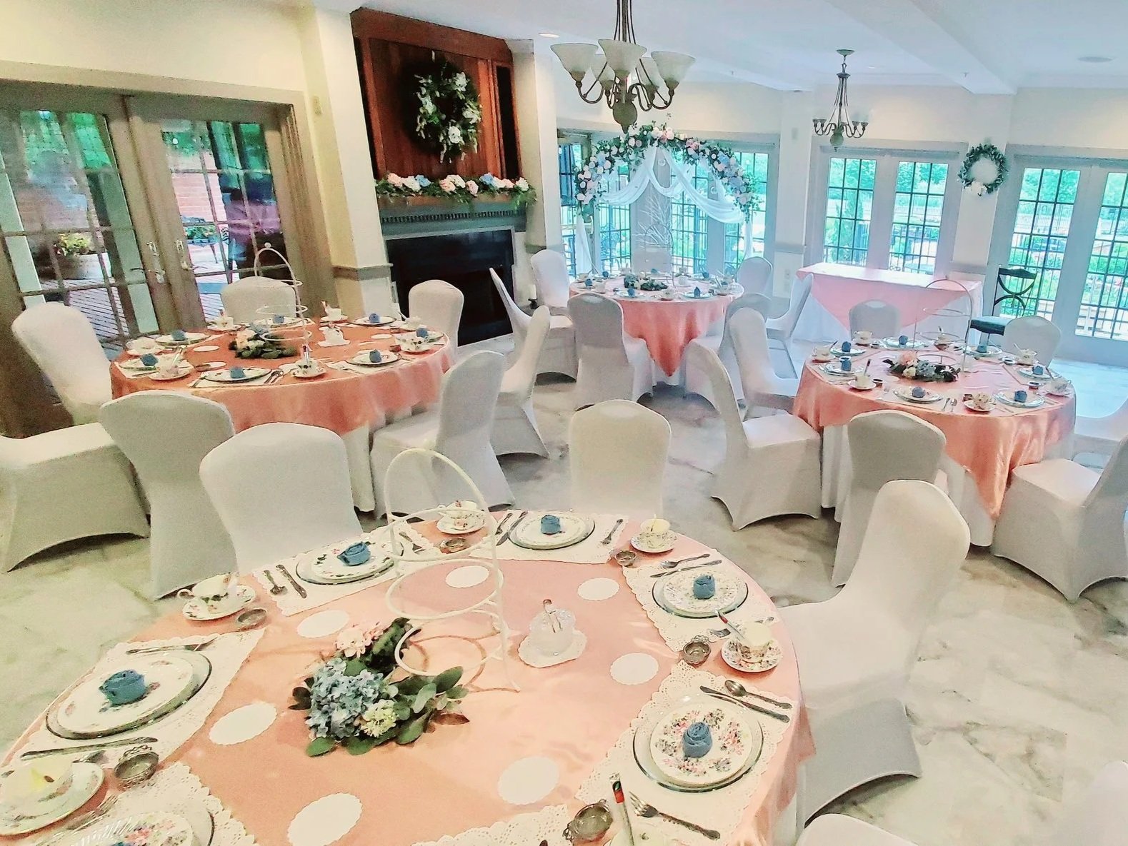 Decorated banquet room with pink tablecloths, white chairs, floral centerpieces, china, and tea sets, set up for a formal event or celebration.